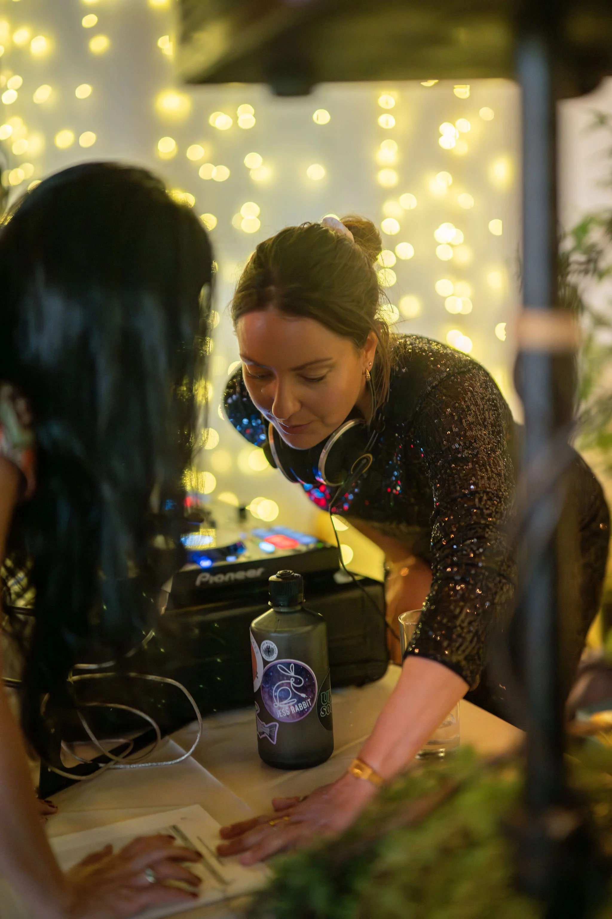 A woman with headphones around her neck leans over a table, focusing on something. She is wearing a black, sparkly top. There are string lights in the background, creating a festive atmosphere. On the table, there is a water bottle, a glass, and some papers.
