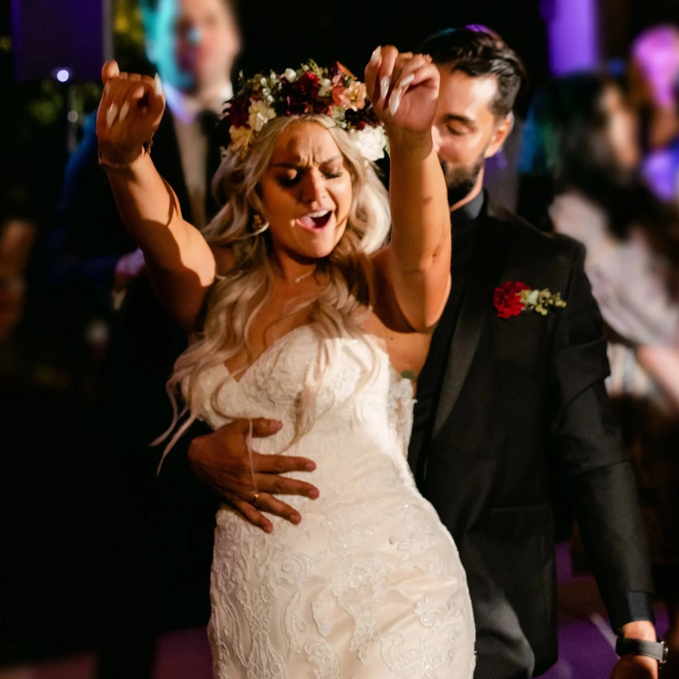A woman with long blonde hair dancing at a wedding, wearing a lace wedding dress and a floral headpiece, with a man in a black suit and floral boutonniere dancing with her.
