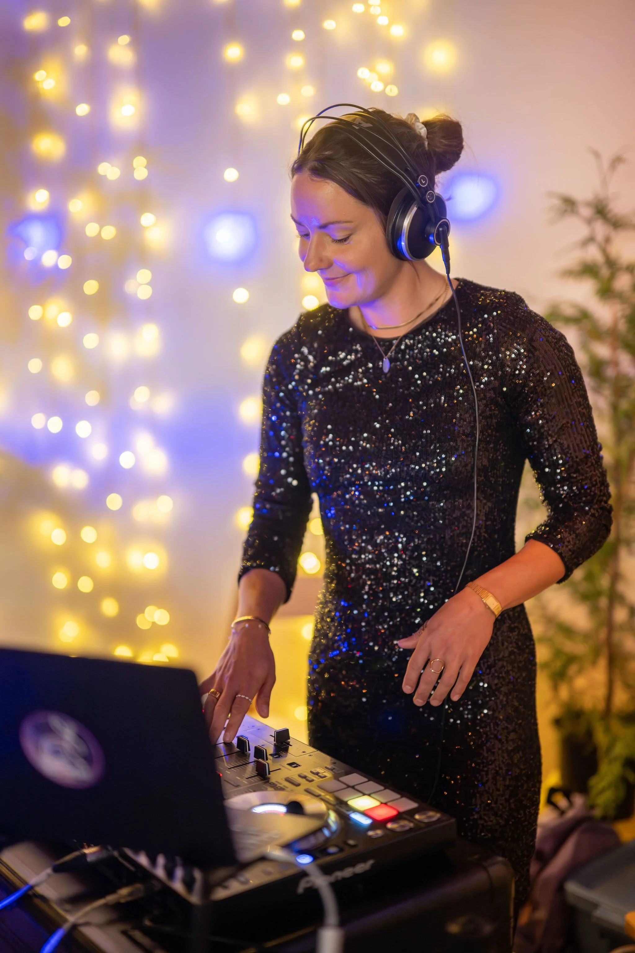 A woman in a black sequined dress DJing at an event, wearing headphones and looking down at her DJ equipment, with yellow string lights in the background.