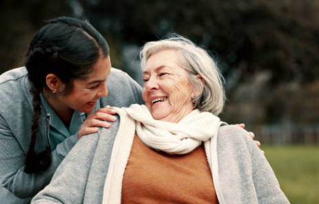 A smiling female nurse with a stethoscope around her neck and a name badge, sitting on a bed with a young boy holding a stuffed animal. The nurse has her arm around the boy, and they appear happy in a bright, modern medical setting.