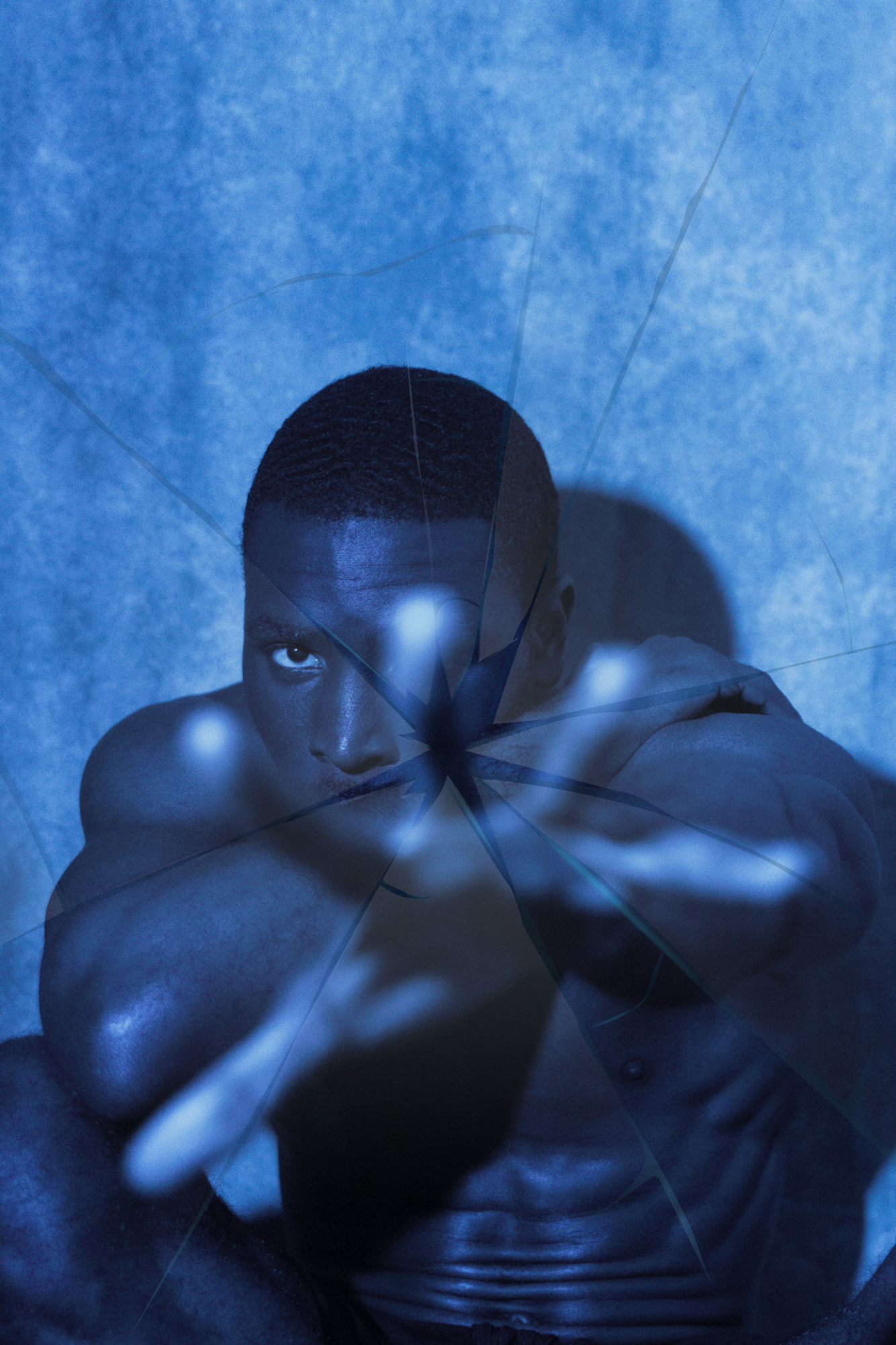 A shirtless man with short hair looking directly at the camera seen through a shattered mirror with blue lighting.