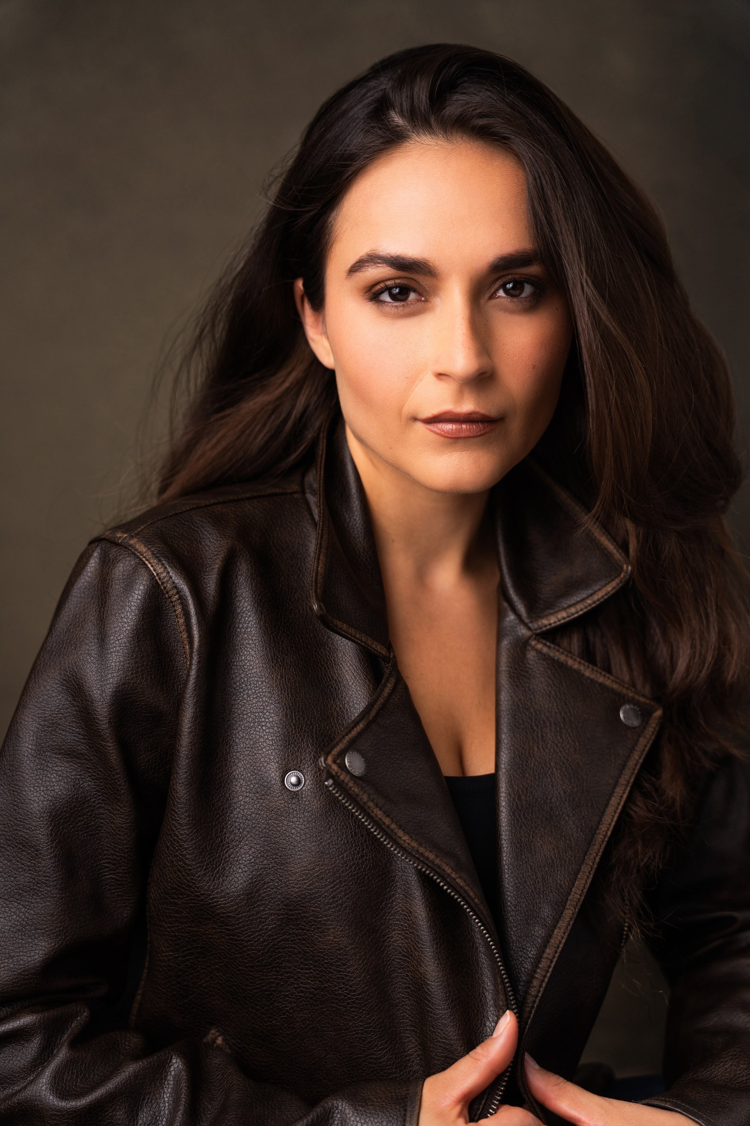 Portrait of a woman with long dark hair wearing a black leather jacket against a dark, neutral background.