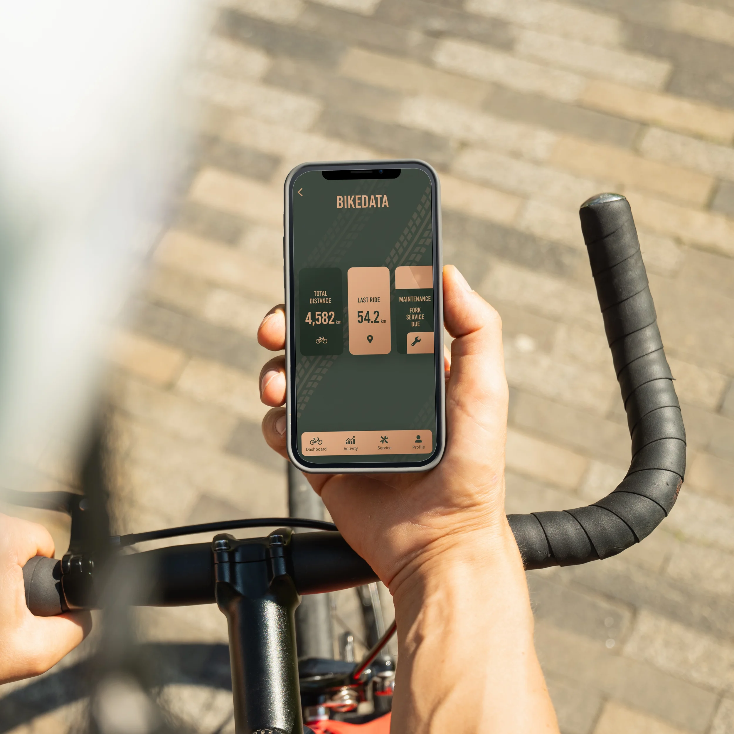 A person holding a smartphone displaying a bike data app while sitting on a bicycle with black handlebars, on a paved surface.