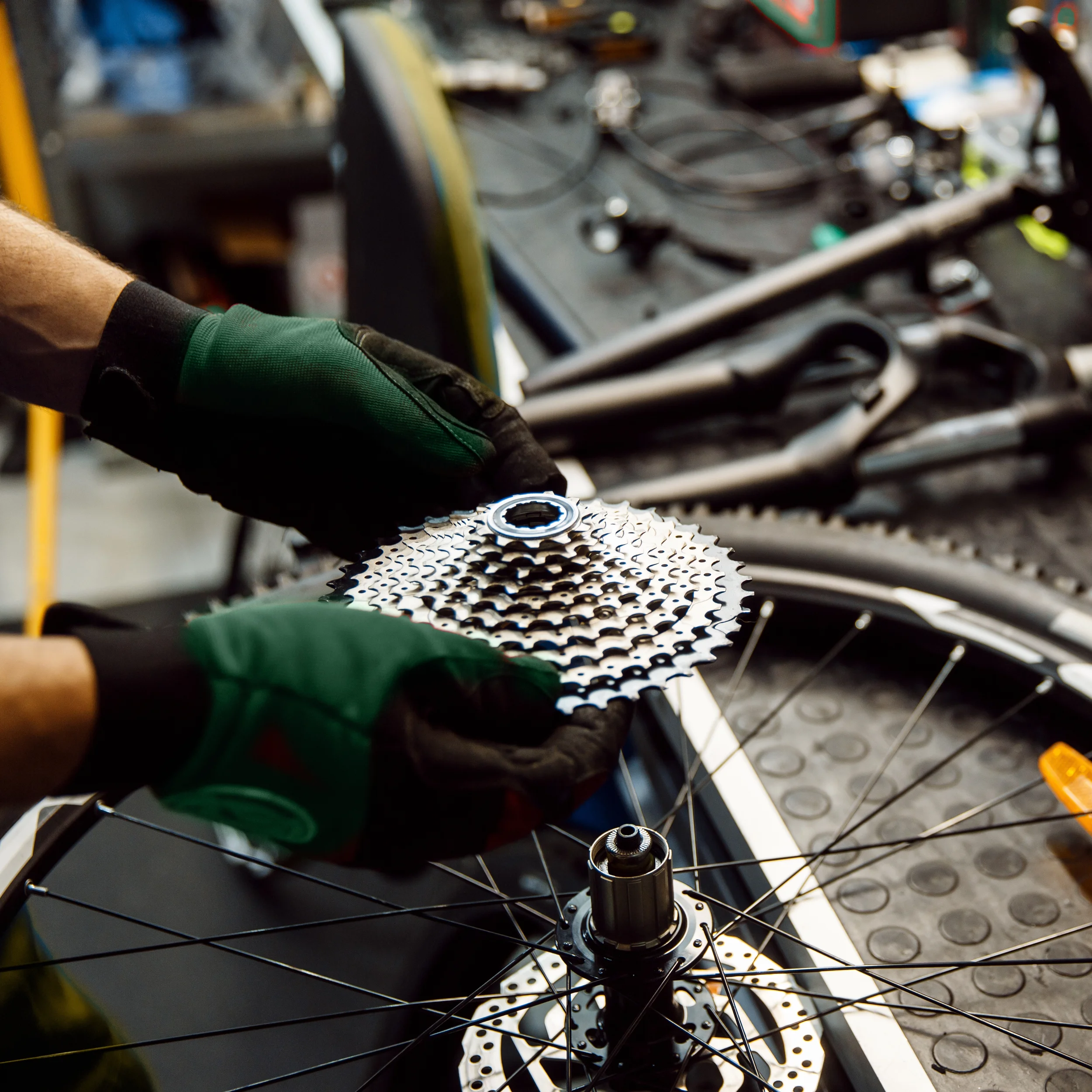 Person wearing gloves assembling a bicycle gear on a workbench, with bicycle wheel and tools in the background.