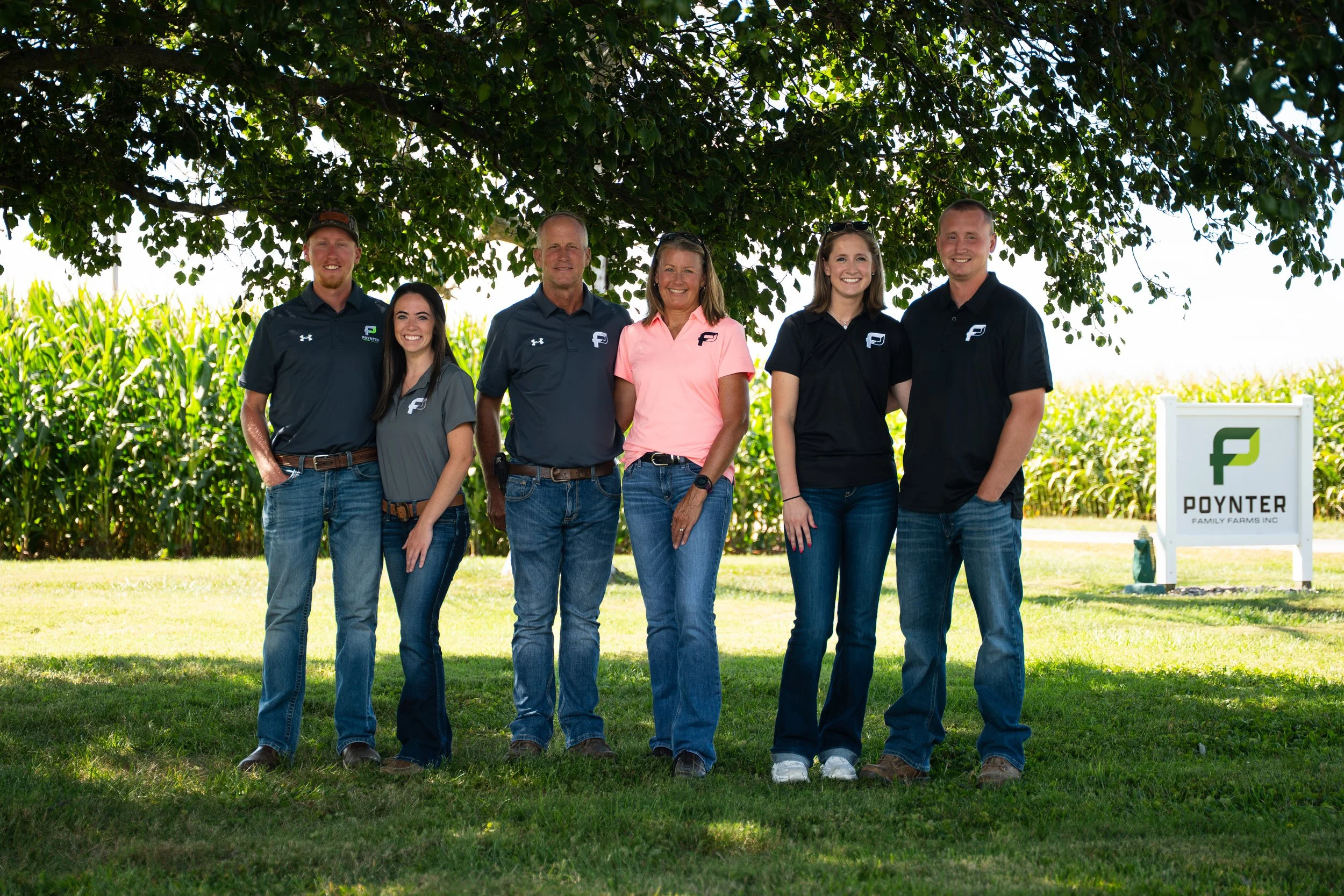 Group of six people standing outdoors beneath a large tree, with a field of corn in the background and a sign that reads "POYNER FAMILY FARMS INC". Poynter Aerial Ag's team.