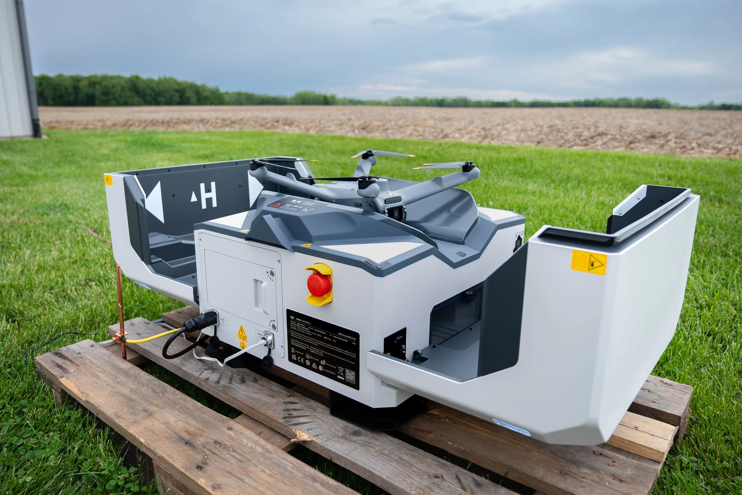 A modern drone with multiple rotors, positioned on a wooden pallet on a grassy field, with farmland and cloudy sky in the background.
