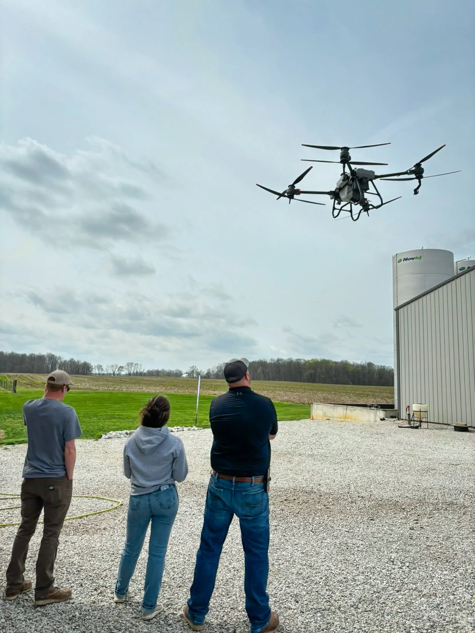 Three people observing a drone flying in the sky near a building with water tanks outdoors.