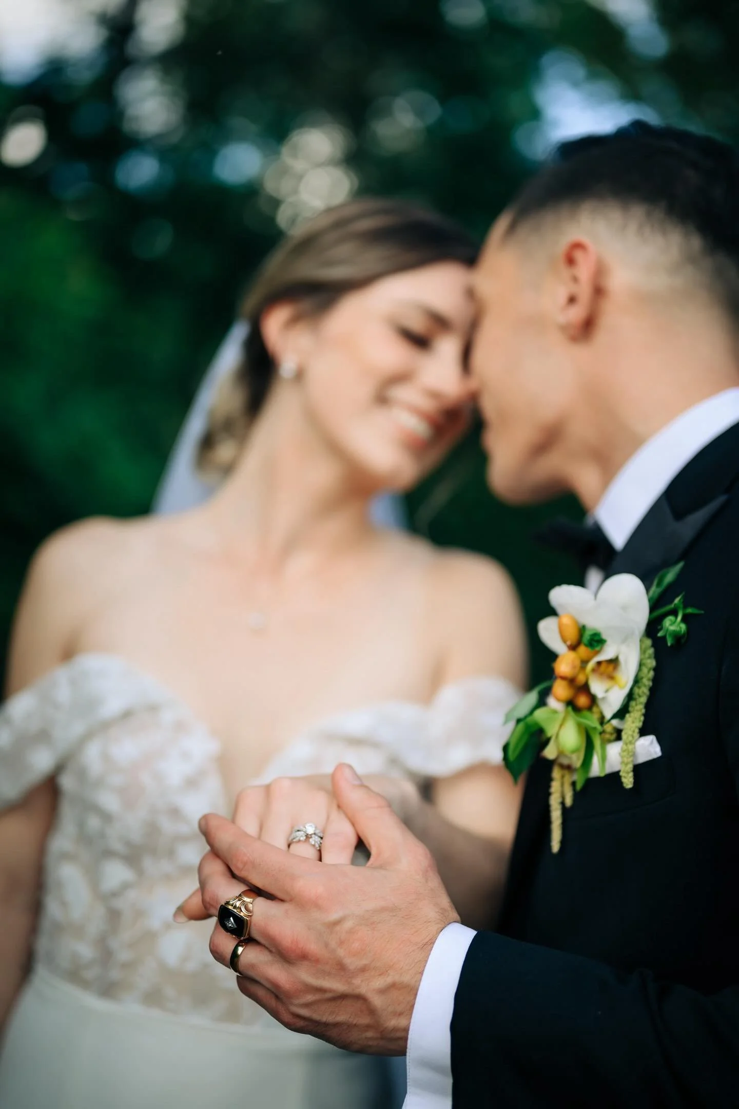 A bride and groom holding hands during their wedding ceremony, showing wedding rings, with bride in a lace dress and groom in a tuxedo with a boutonniere, outdoors with greenery in the background.
