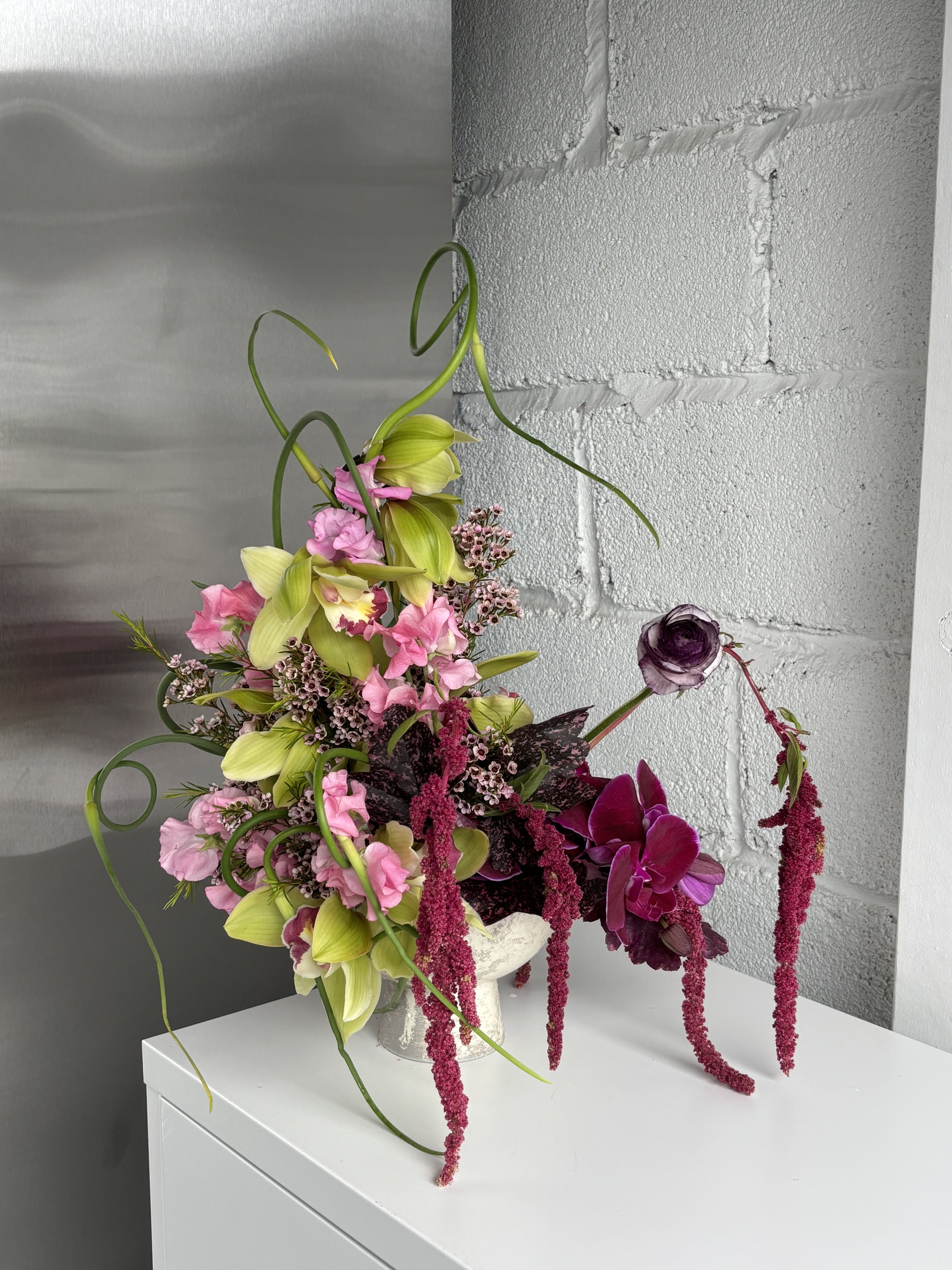 A floral arrangement featuring pink, purple, and green flowers in a white vase on a white surface against a background of a light gray brick wall and a metallic panel.