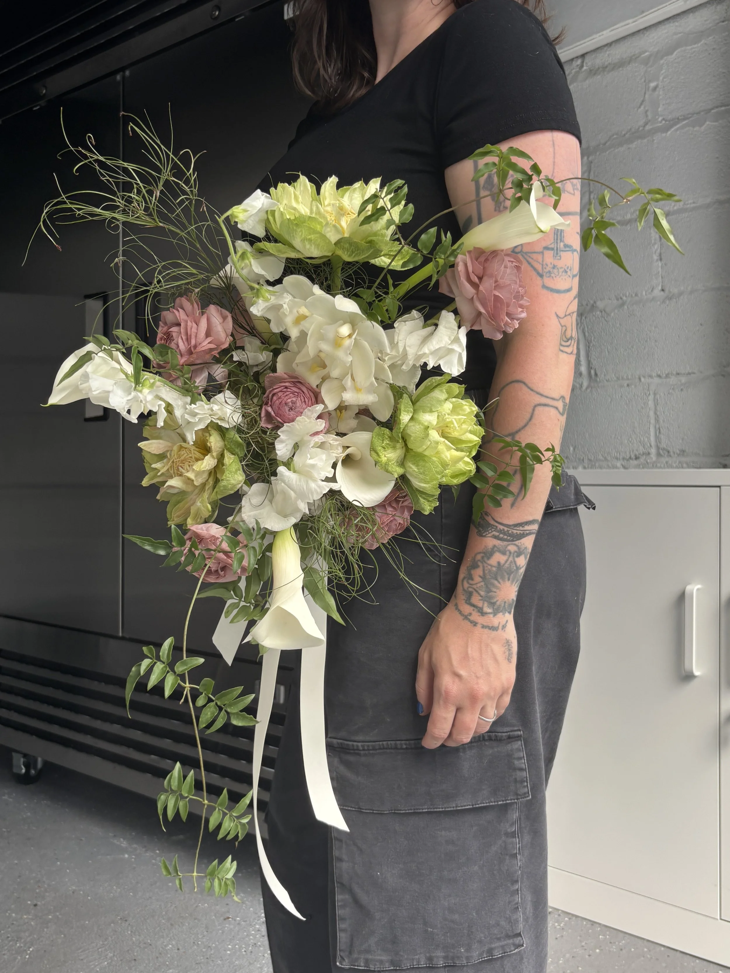 Person holding a bouquet of white and pink flowers with tattoos on their arms, wearing a black shirt and gray pants.