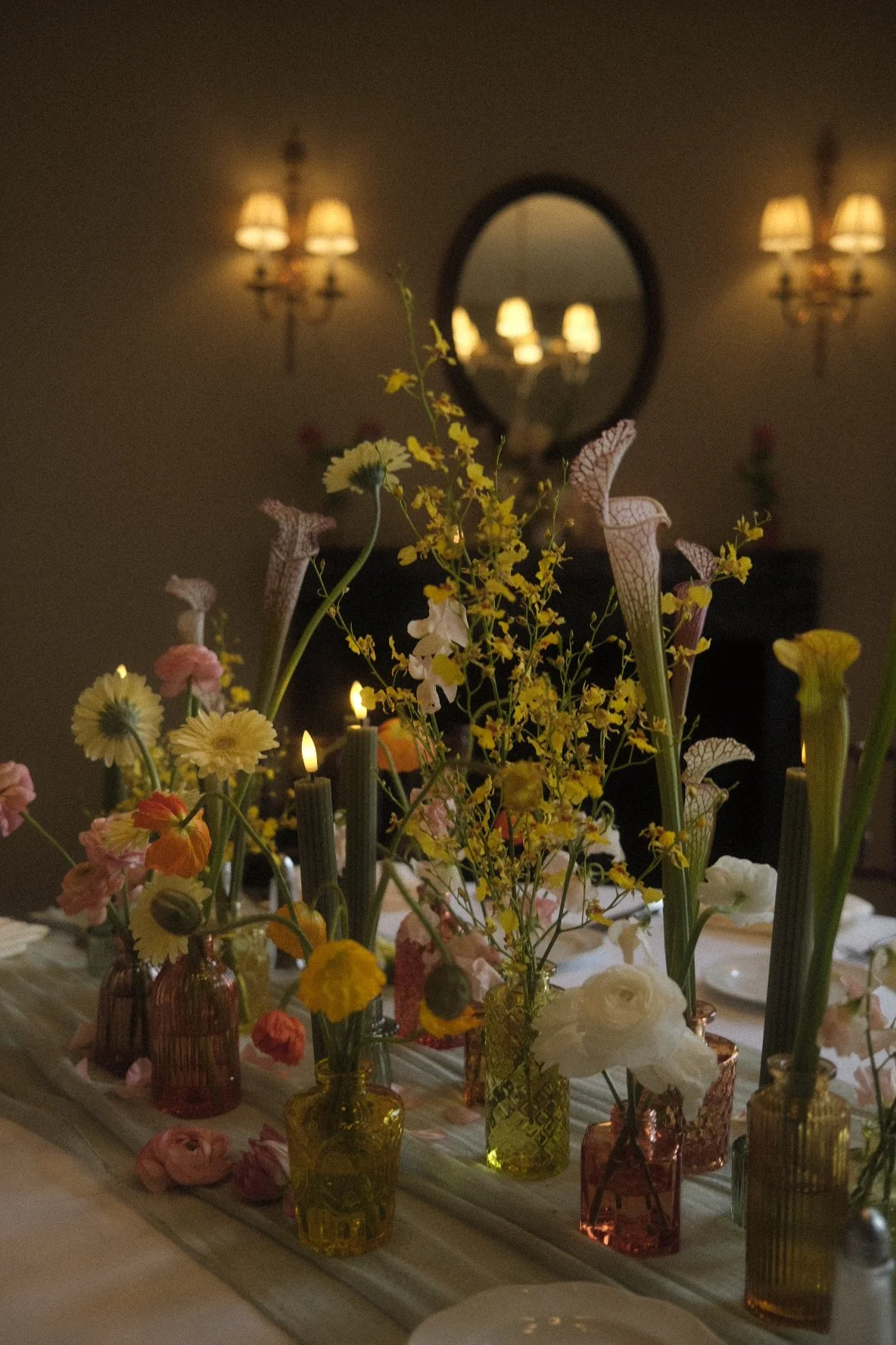 Elegant table centerpiece with various colorful flowers in small glass vases on a white cloth table, with candles and a mirror reflecting wall sconces in the background.