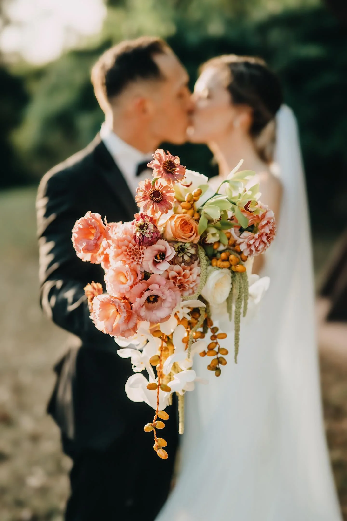 A couple kissing in wedding attire with a colorful bouquet of flowers in focus in the foreground, outdoors with blurred greenery background.
