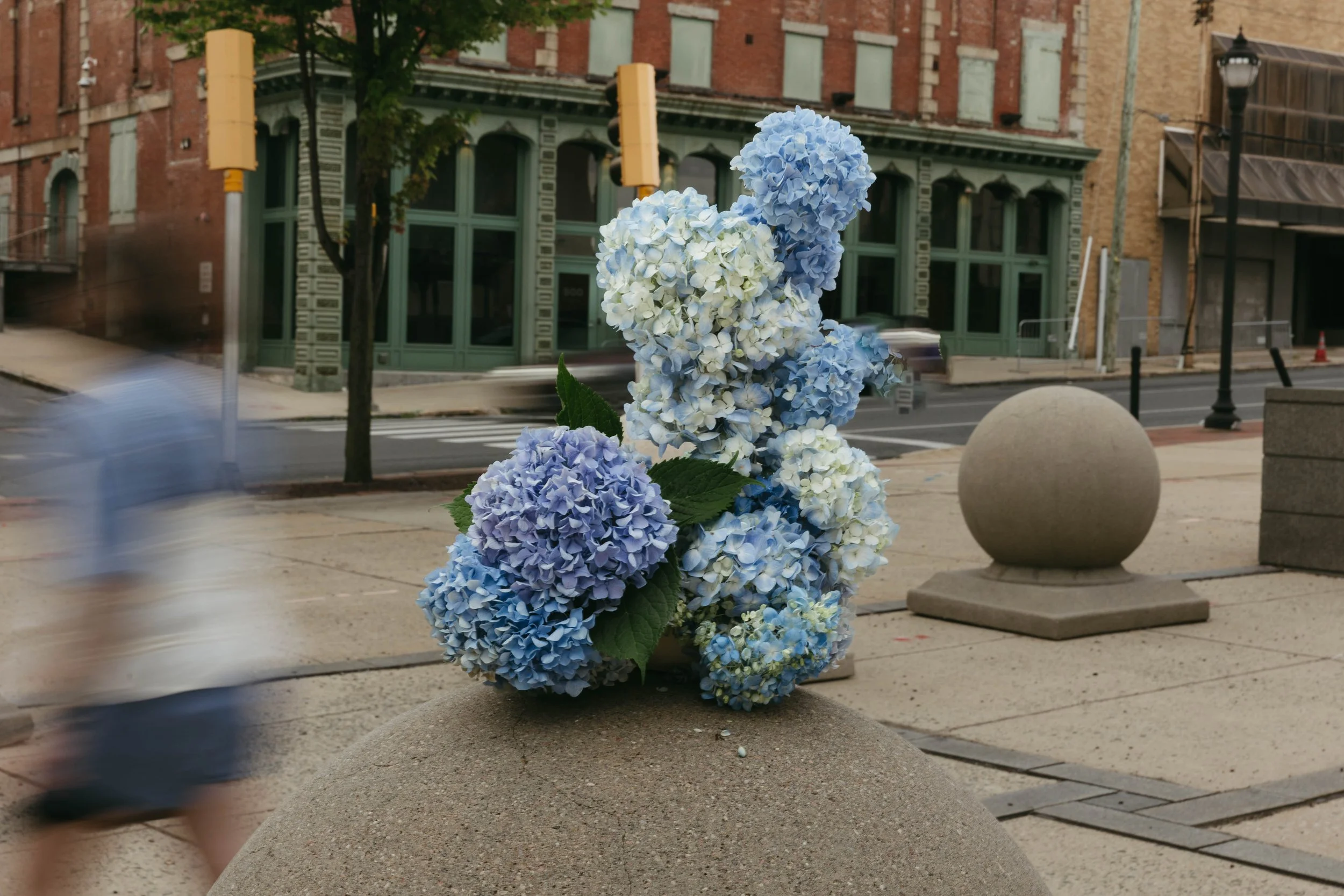 A cluster of blue and white hydrangea flowers arranged on a concrete base on a city sidewalk, with blurry pedestrian walking by and an urban street scene in the background.
