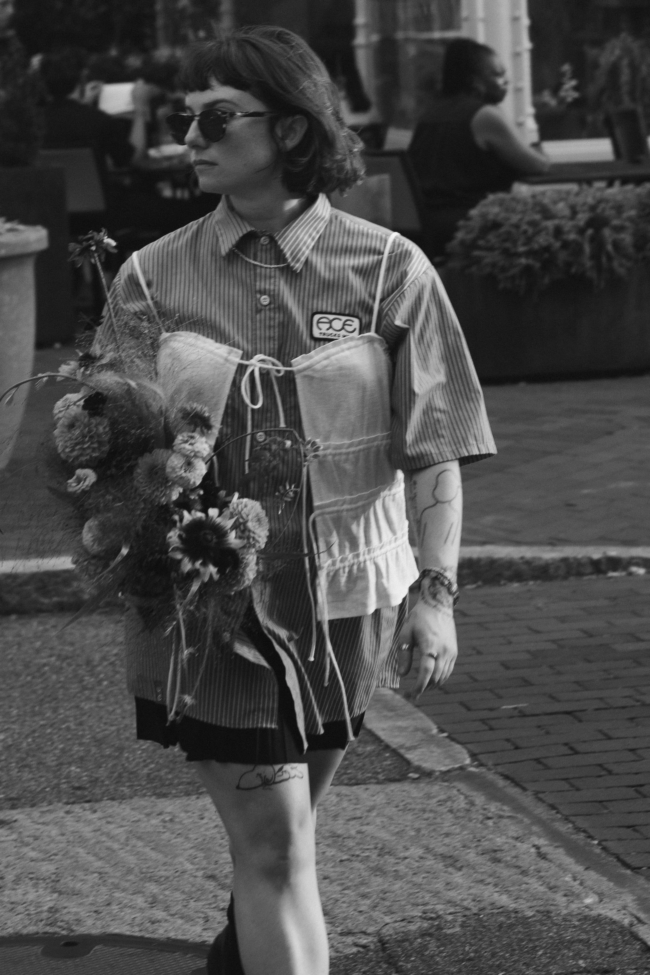 A woman in a striped shirt and apron walking outdoors while holding a bouquet of flowers, with other people sitting at tables in the background.