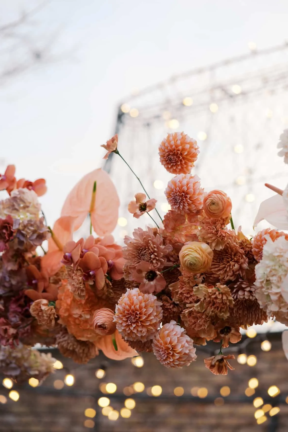 Close-up of pink and peach flowers, including dahlias, roses, and anthuriums, arranged on a table. In the background, there are blurred lights creating a bokeh effect, suggesting a festive or decorative setting.