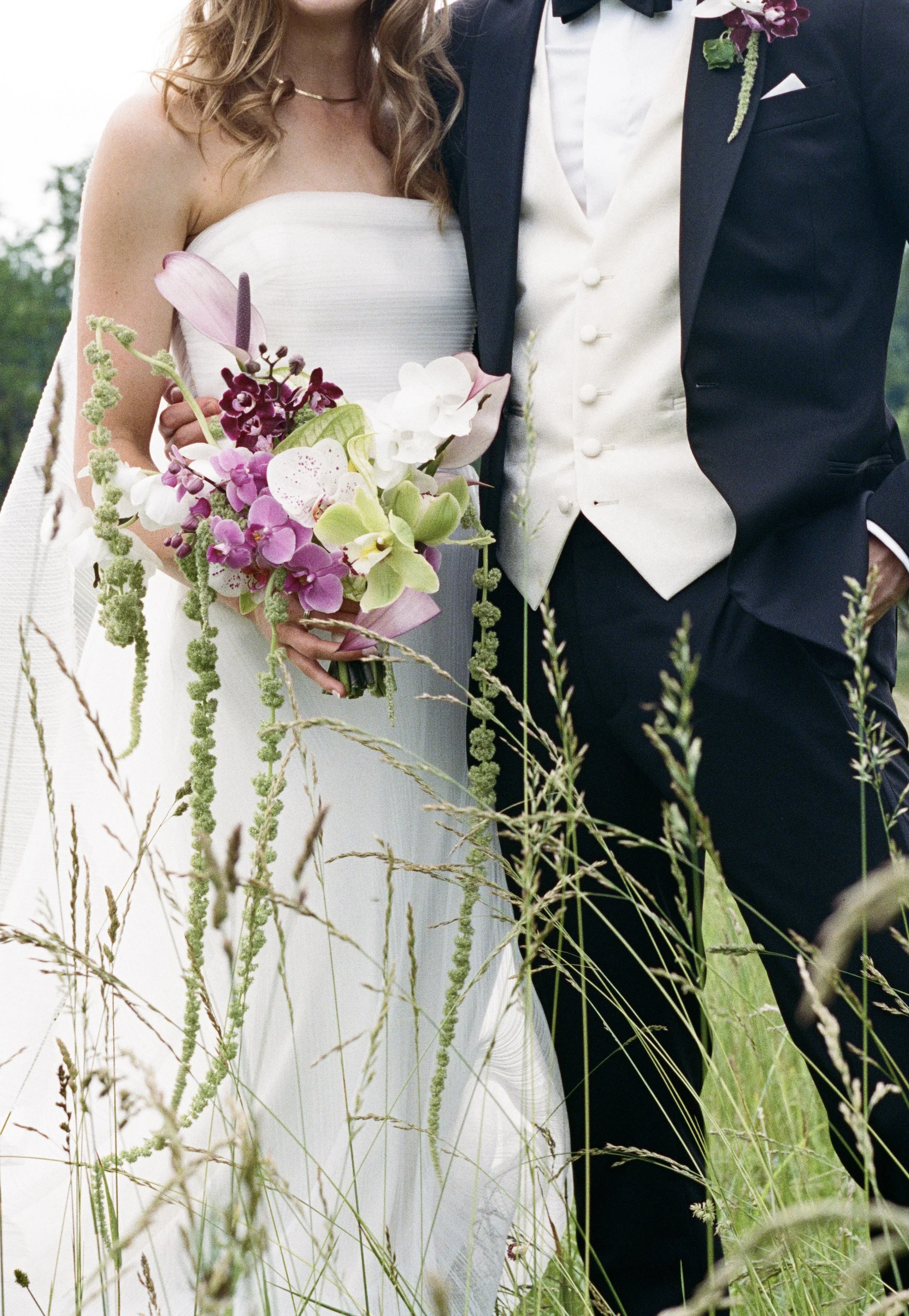 A bride and groom standing together outdoors, with the bride holding a bouquet of pink, purple, and white flowers amidst tall grass.