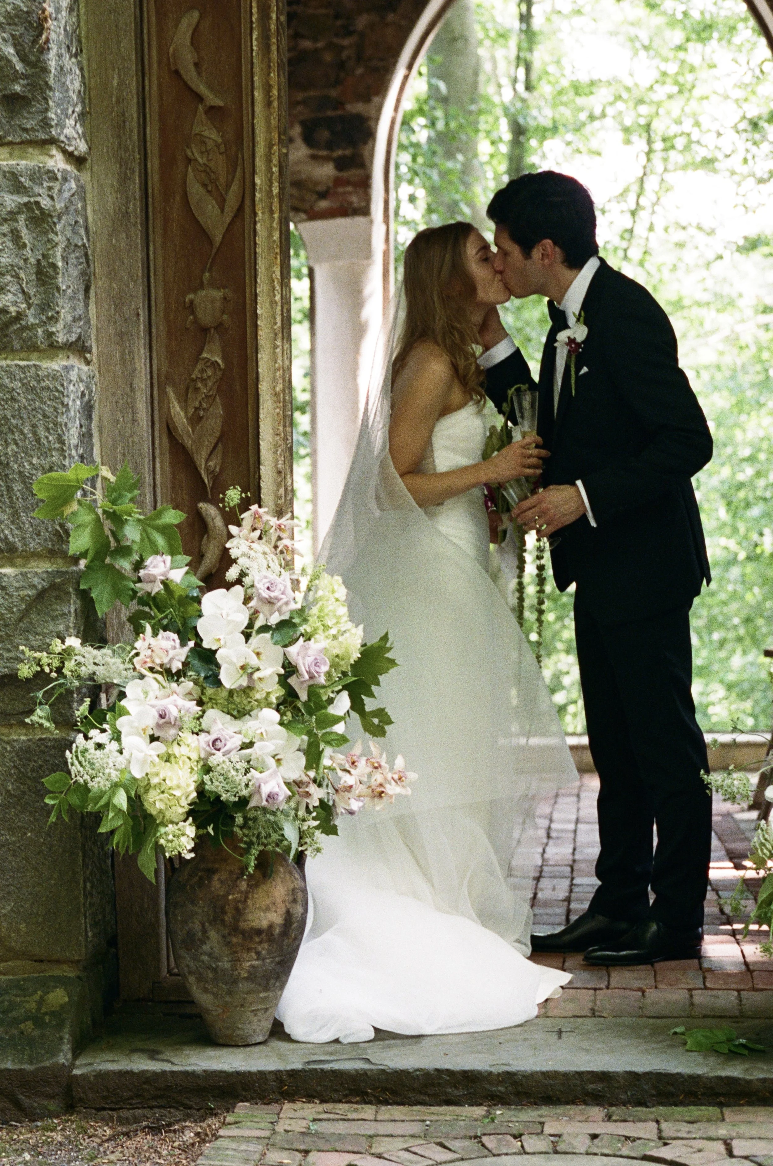 A bride and groom kissing during their wedding ceremony outdoors under a stone archway, with a floral arrangement nearby.