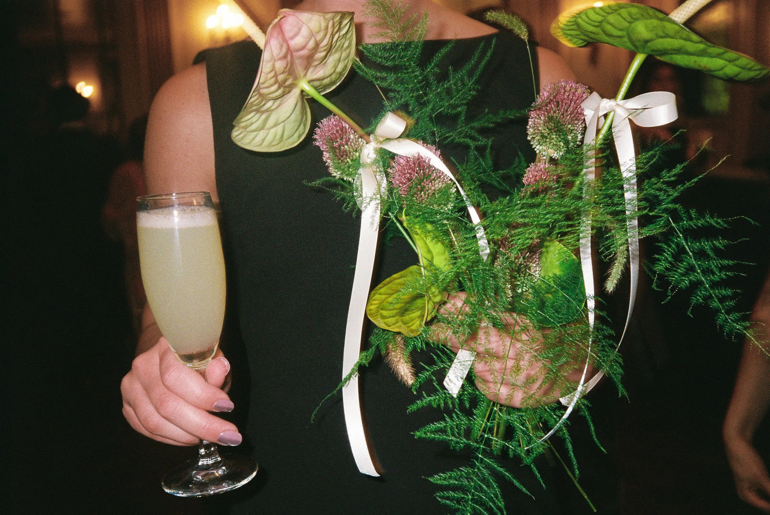 Man holding a champagne glass with a floral arrangement on his chest. The arrangement includes large green leaves, pink flowers, and ferns, with a white ribbon. The man is wearing a black outfit and appears to be at an indoor celebration or event.