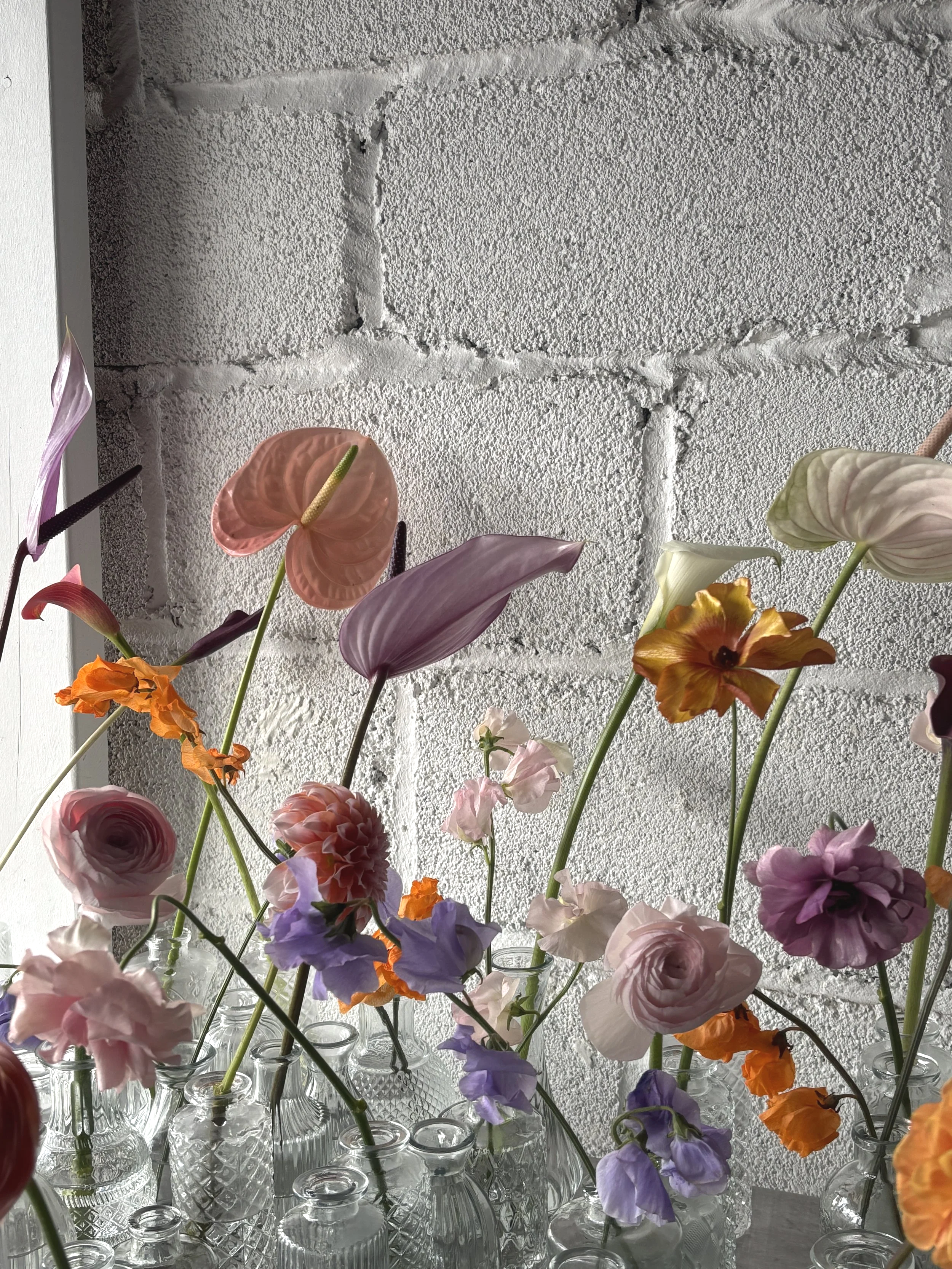 Various colorful flowers in glass vases against a white brick wall.