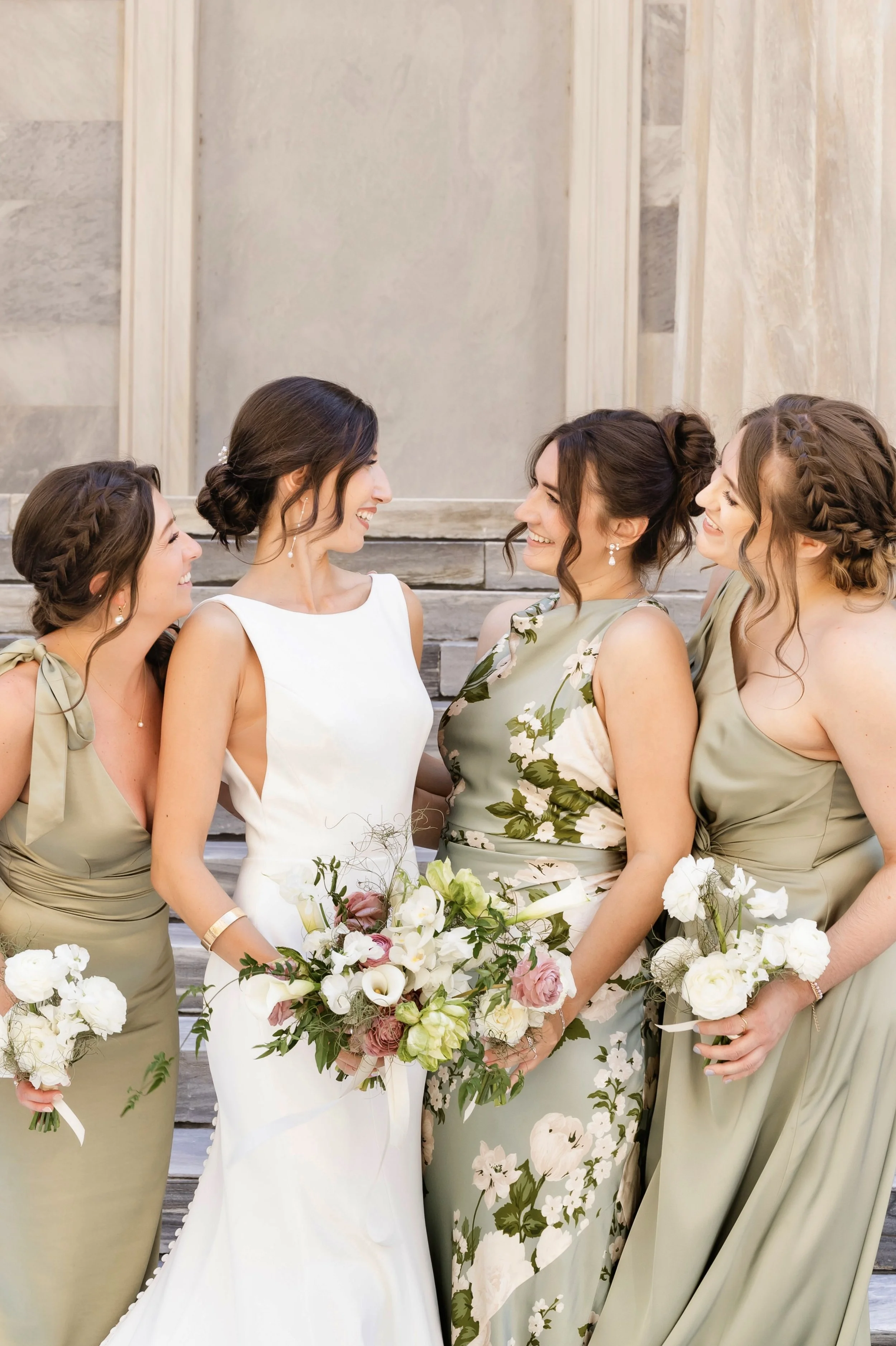 A bride and four bridesmaids smiling and looking at each other outside on steps, holding bouquets of white, pink, and green flowers.
