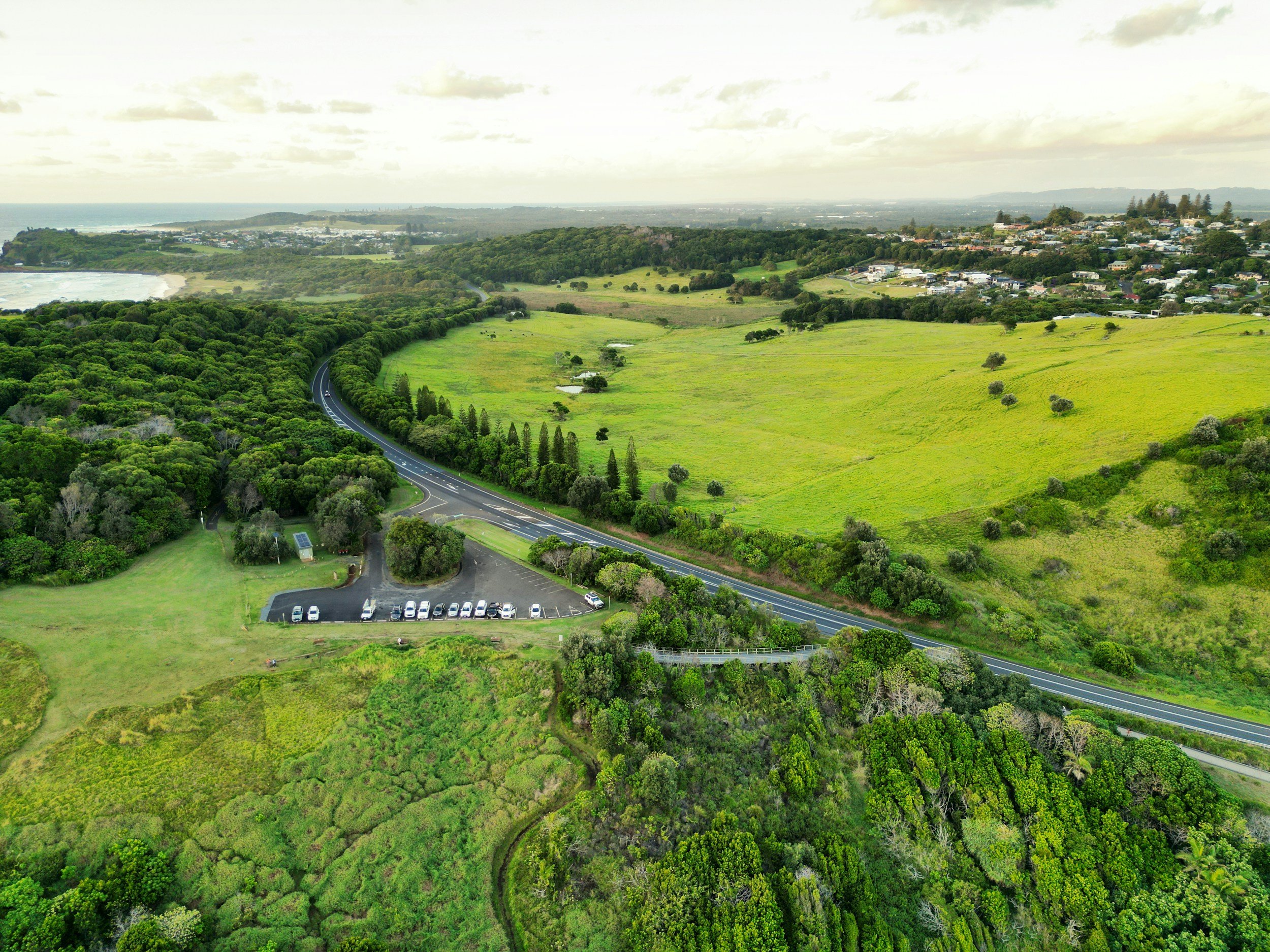 Aerial view of a winding road through green hills and trees near a parking lot with cars, with a small town and ocean in the distance under cloudy skies.