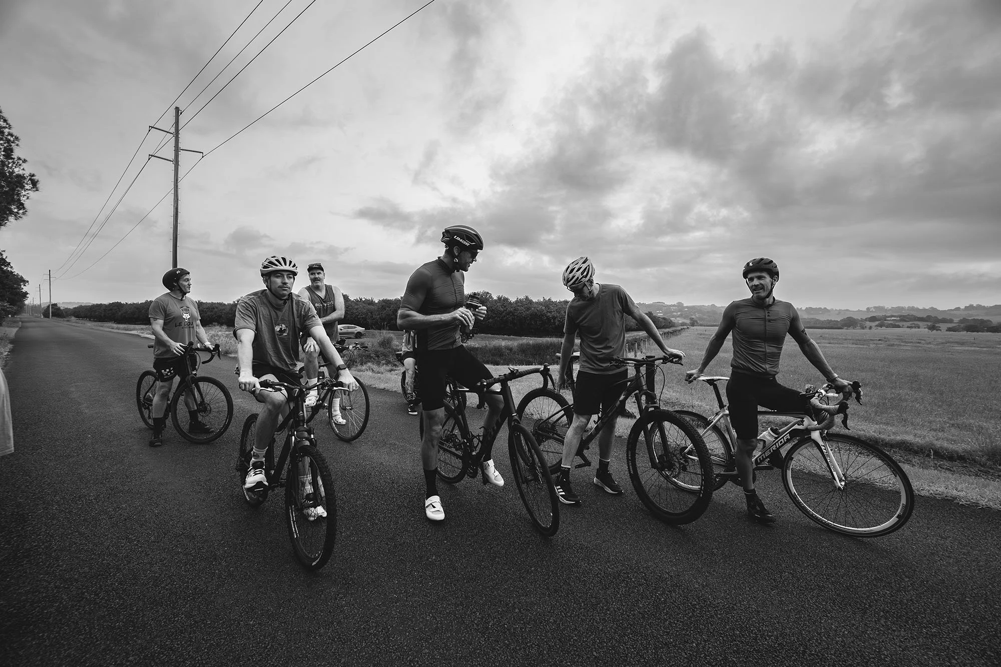 A group of six men on bicycles standing on a paved road in a rural area with open fields and cloudy sky in the background, some talking and one looking at his phone.