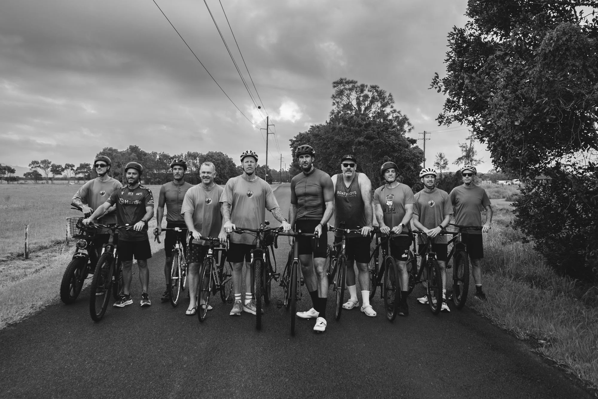 A group of nine men with bicycles standing on a rural road during cloudy weather, wearing casual and athletic clothing including helmets.