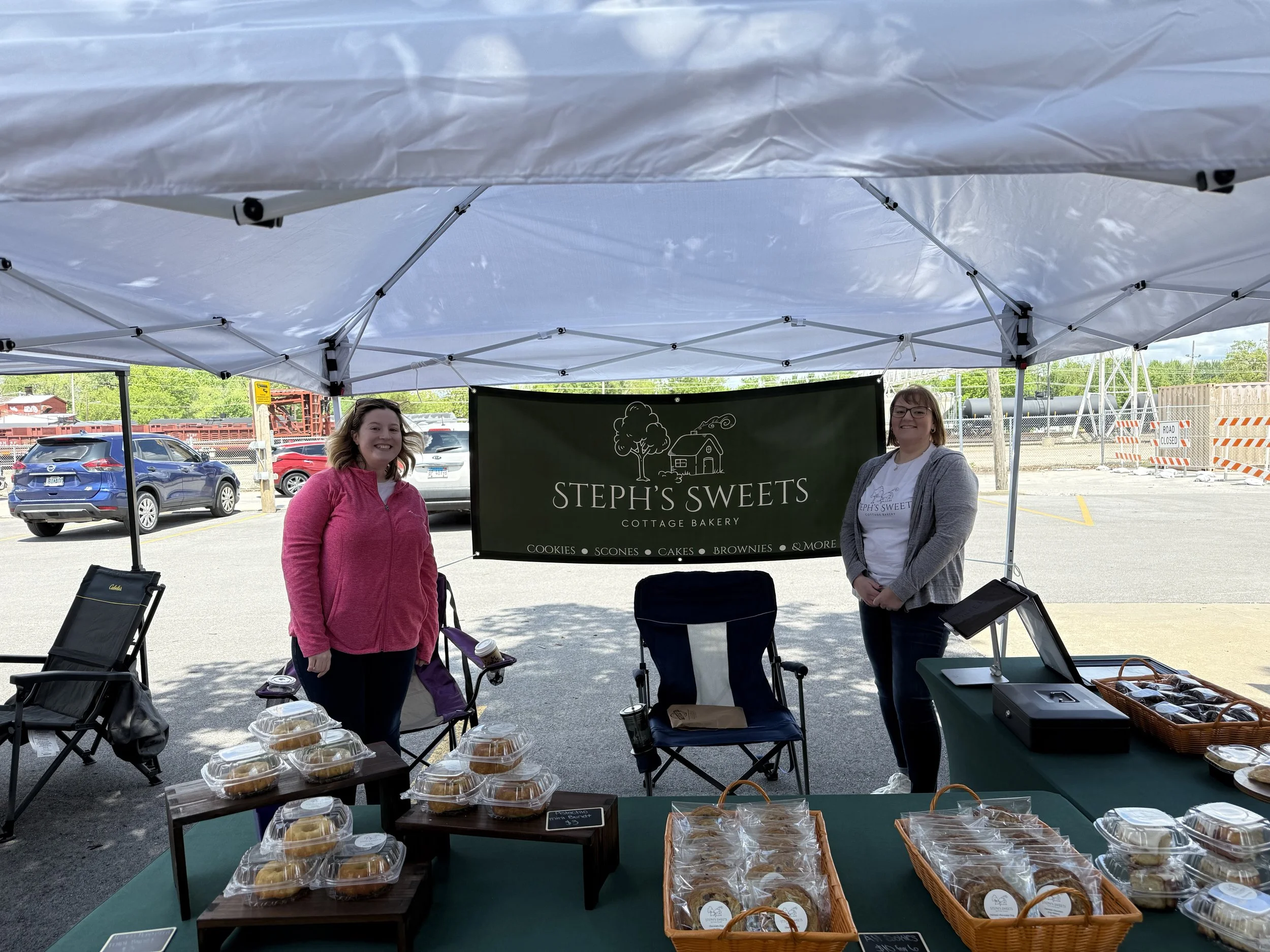 Two women standing behind a table of baked goods at an outdoor market stall called Steph's Sweets. The table displays cookies, scones, and cakes in plastic containers, with a green sign behind them on a white tent. Cars are parked in the background.