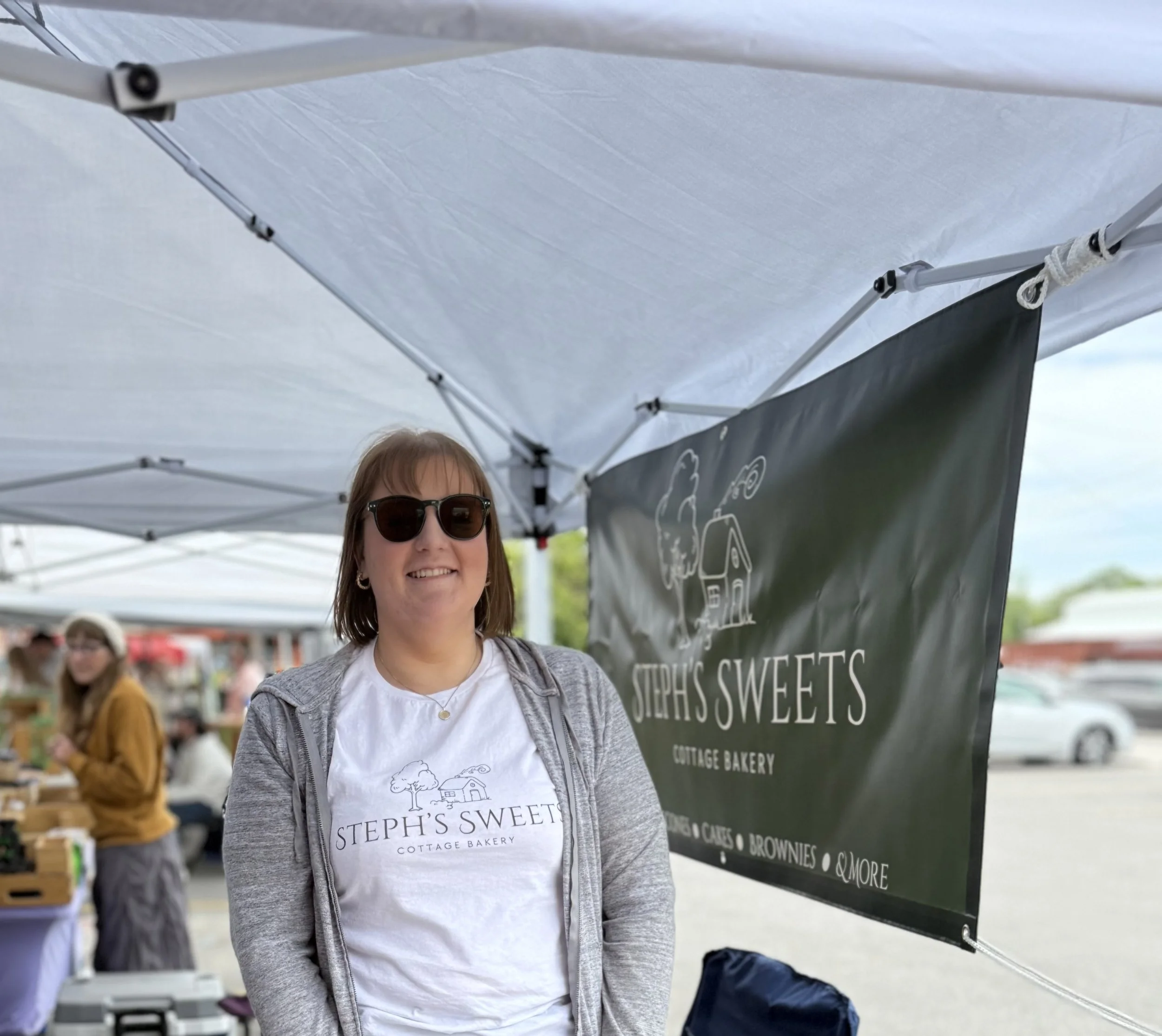 A woman smiling at a market booth under a white canopy, wearing sunglasses and a white t-shirt with a bakery logo, with other vendors and customers in the background.