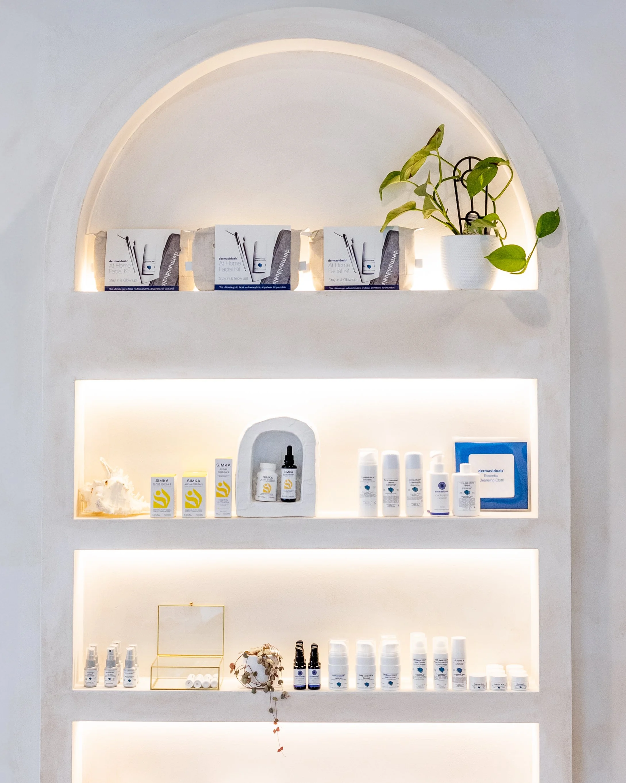 Display shelves with skincare and beauty products, a white potted plant, and decorative items in a minimal-tinted room.