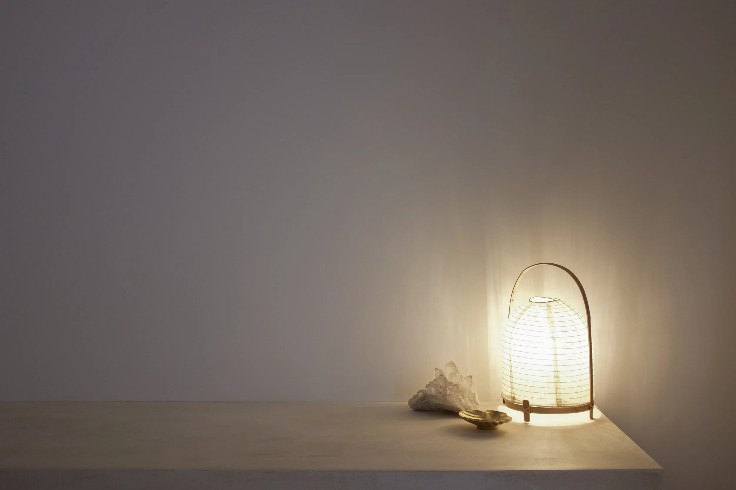 A lit paper lantern, a cluster of clear quartz crystals, and a small beige object on a light-colored tabletop against a plain wall.