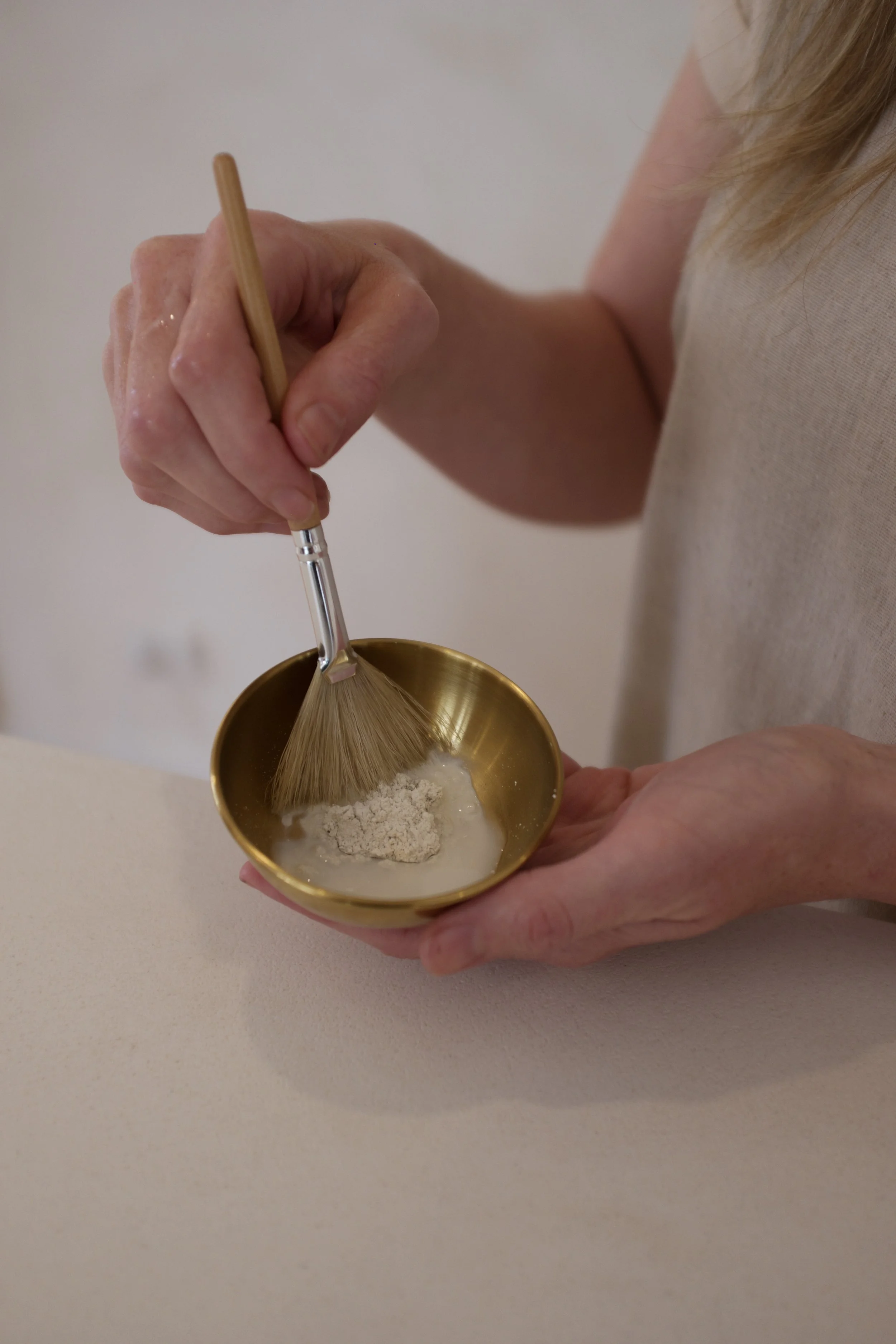 Person mixing a powder and liquid in a gold bowl using a small whisk.