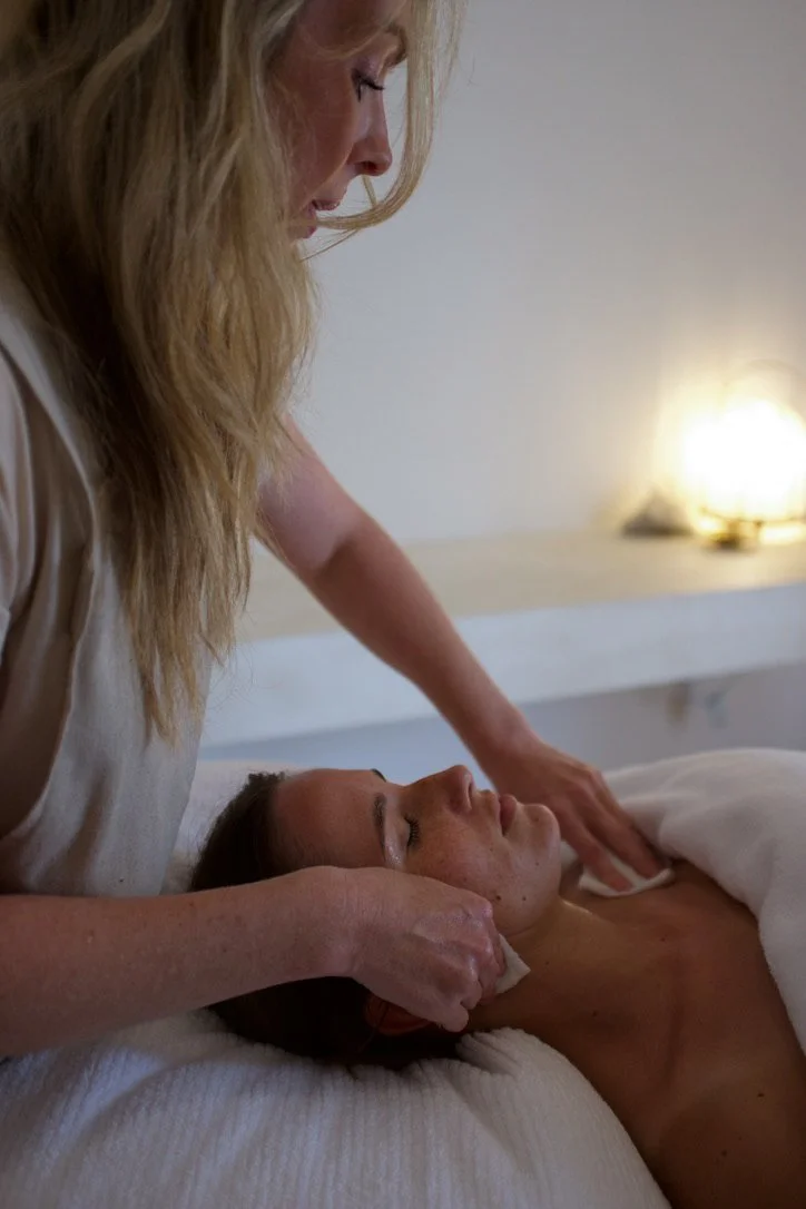 A woman receiving a facial massage while lying on a bed, with another woman gently massaging her face in a spa setting.