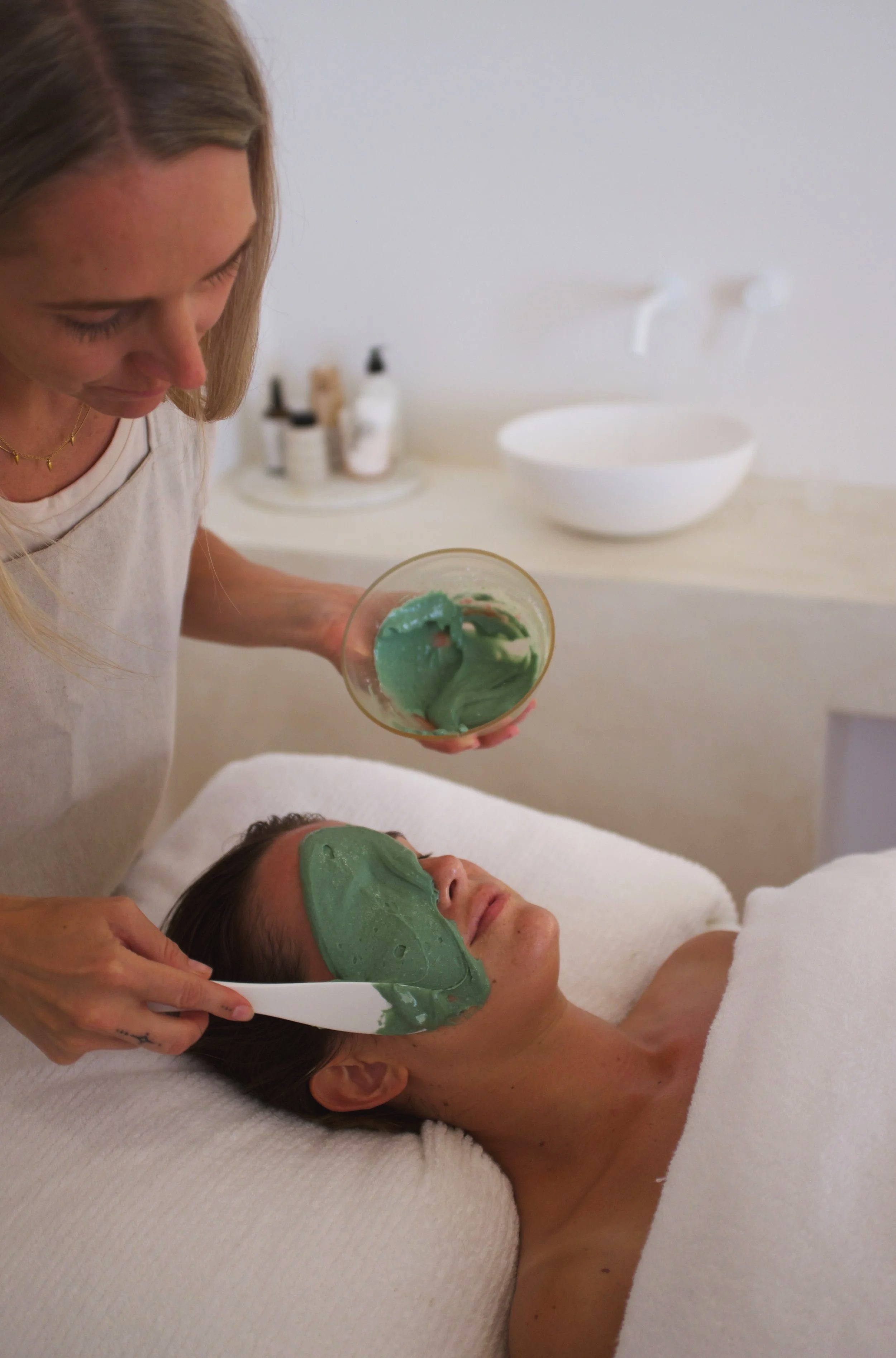 A woman is applying a green facial mask on a woman lying on a treatment bed in a spa or skincare clinic.
