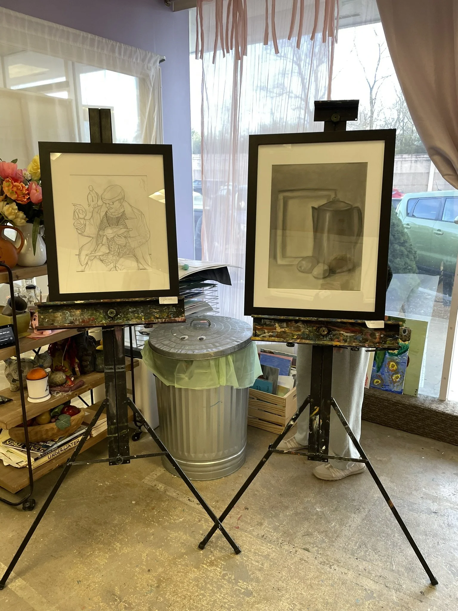 Two framed drawings on easels, one depicting a person reading a magazine and the other a still life with a teapot and stones, in an art studio with a window and various art supplies.
