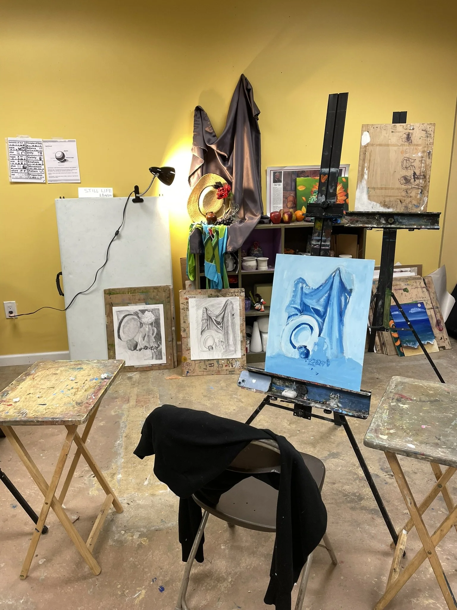 Art studio with paintings on the wall, an easel with a blue painting, and various art supplies and sketches. Two wooden tables with paint splatters, a chair with a black jacket, and a yellow wall with a lamp illuminating the space.