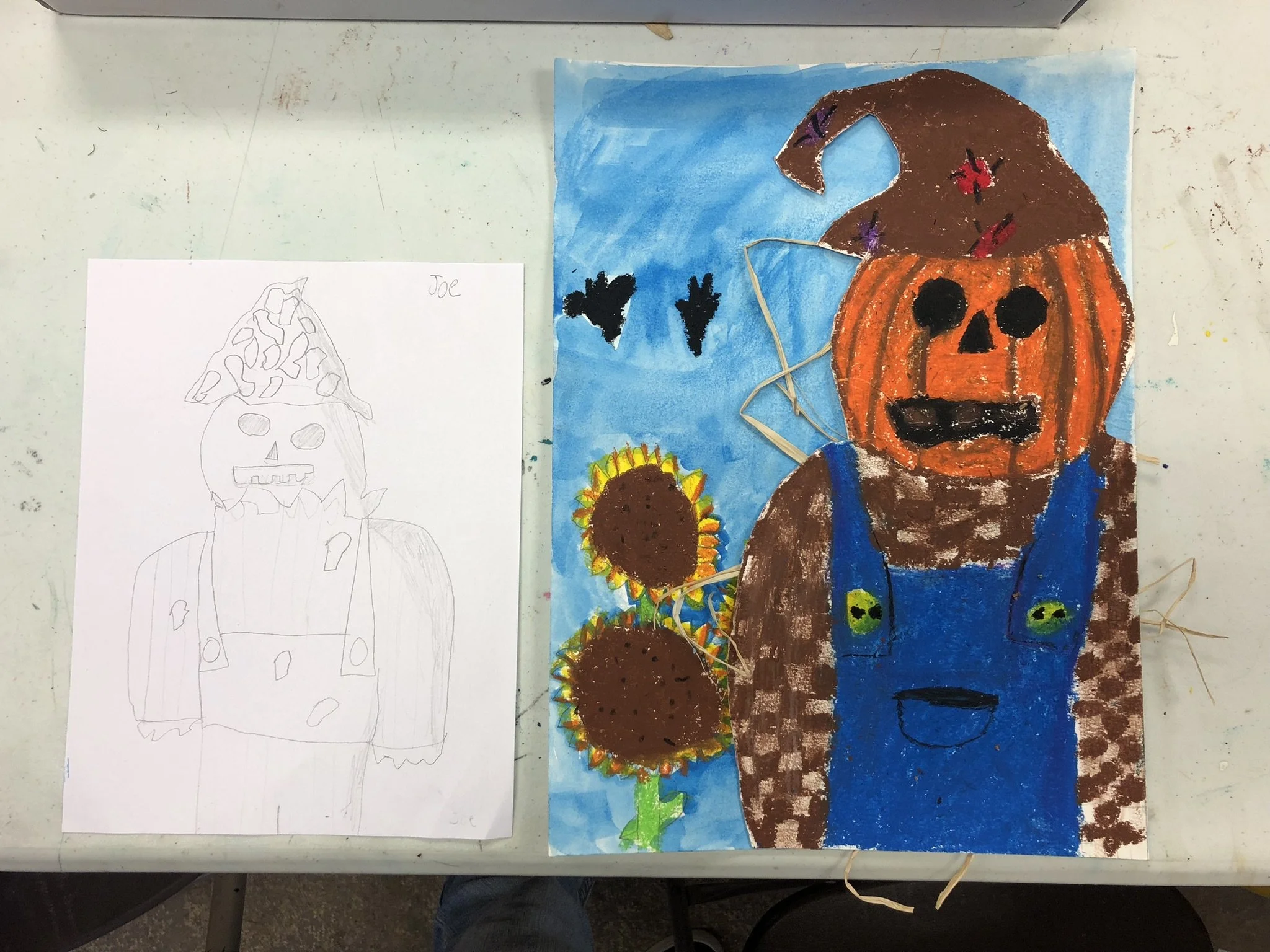 Child's Halloween art project featuring a pumpkin-headed character with a brown hat, black eyes, and a blue outfit, alongside two sunflowers, against a blue background with black bat shapes.