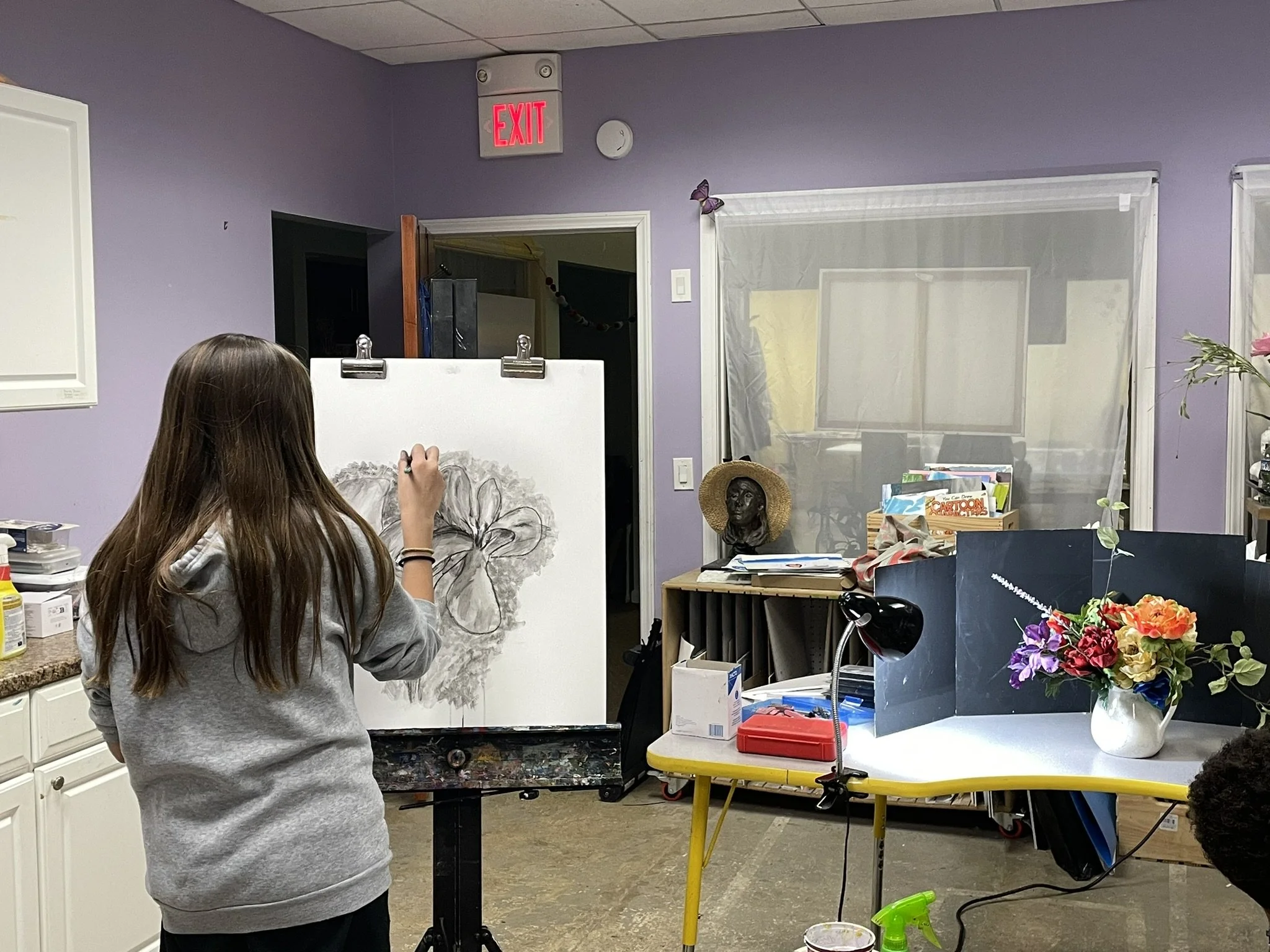 A girl drawing a flower on a canvas in an art classroom with purple walls, a table with art supplies, and decorated with flowers and artwork.