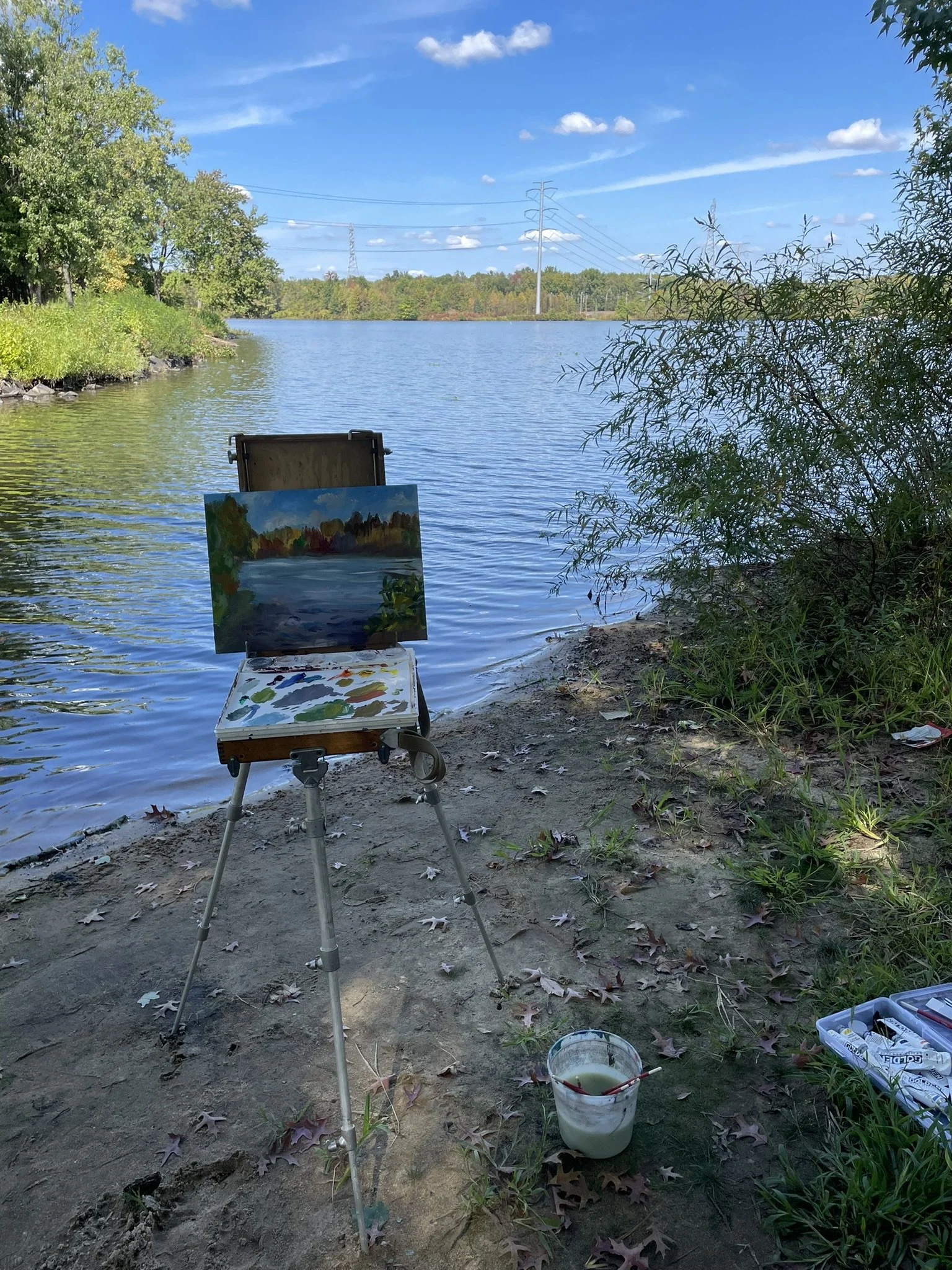 A painter's setup by a lakeside with a canvas on an easel, depicting a landscape scene with water and trees, under a blue sky with some clouds.