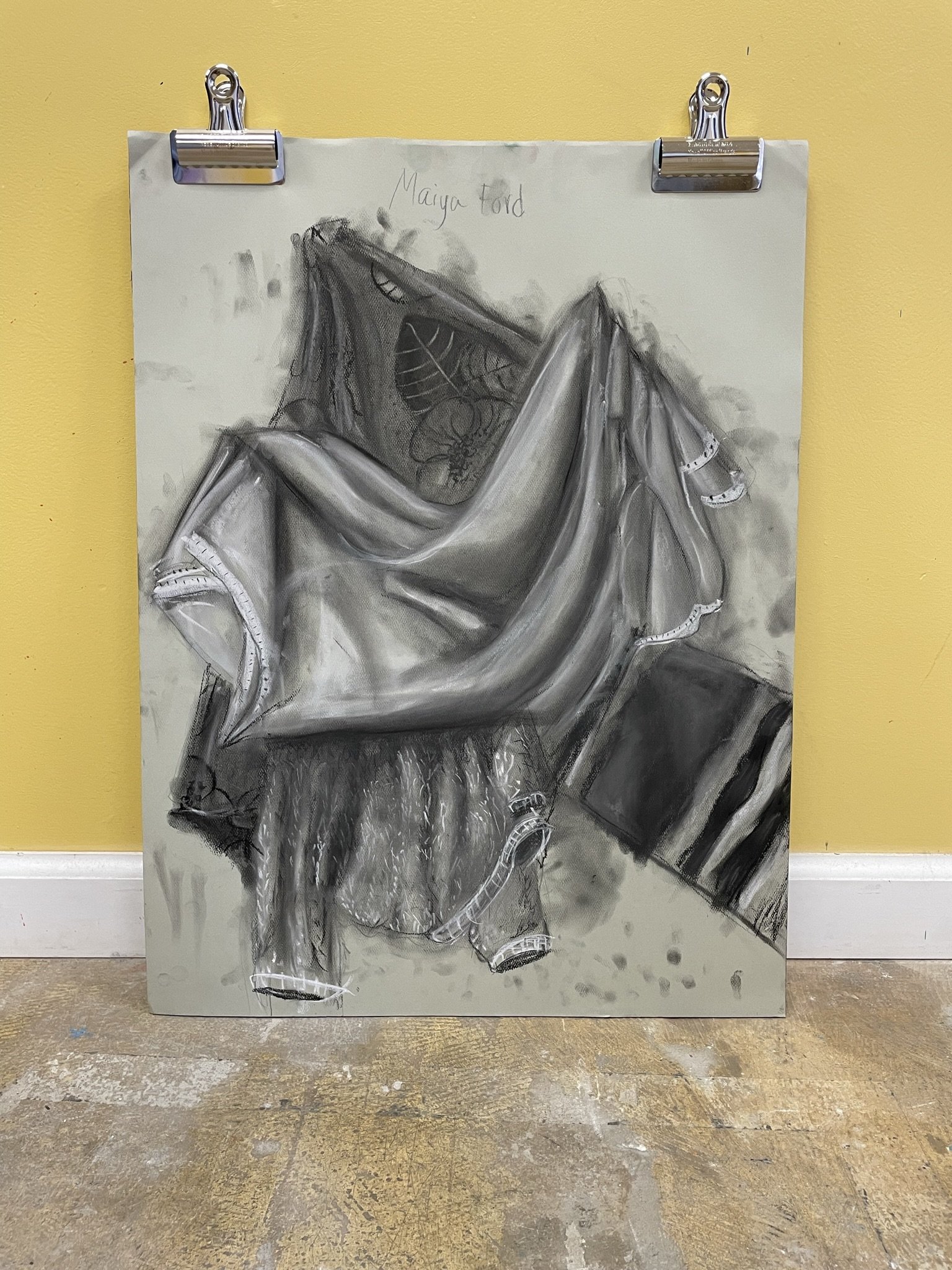 A grayscale drawing of a person sitting with legs crossed, draped in a cloth, with detailed tattoos or artwork on the upper body, on paper attached with metal clips against a yellow wall.