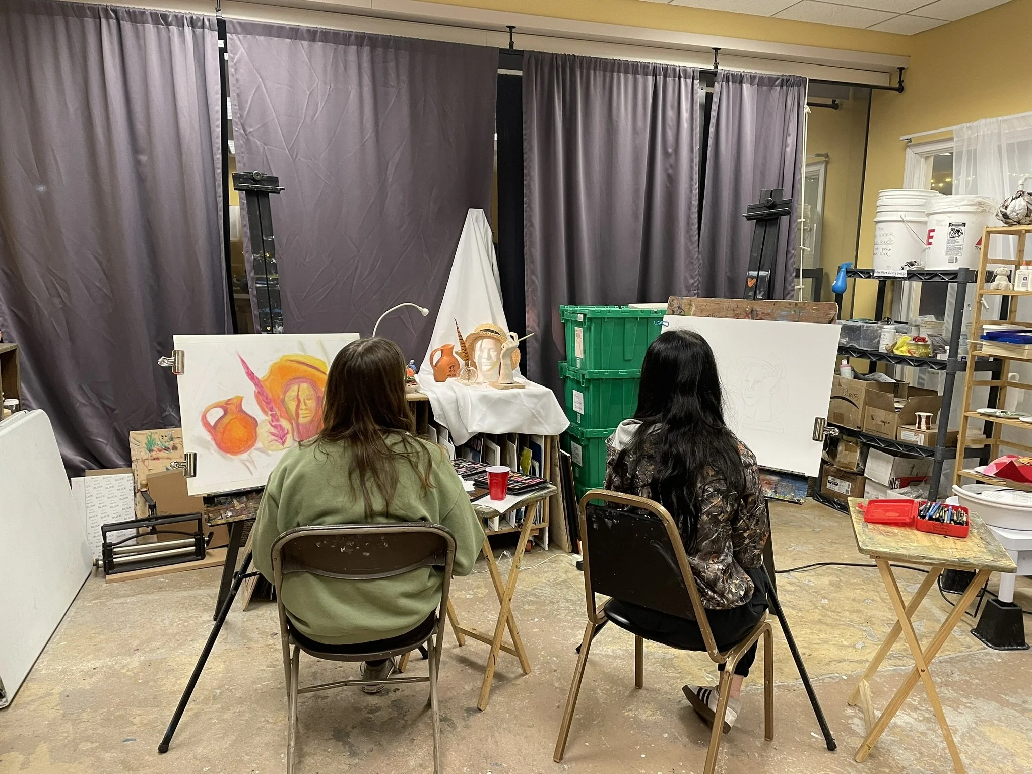 Two women sit in chairs facing an art setup with canvases, supplies, and sculptures in an art studio or classroom, with a grey curtain behind and various art materials around.