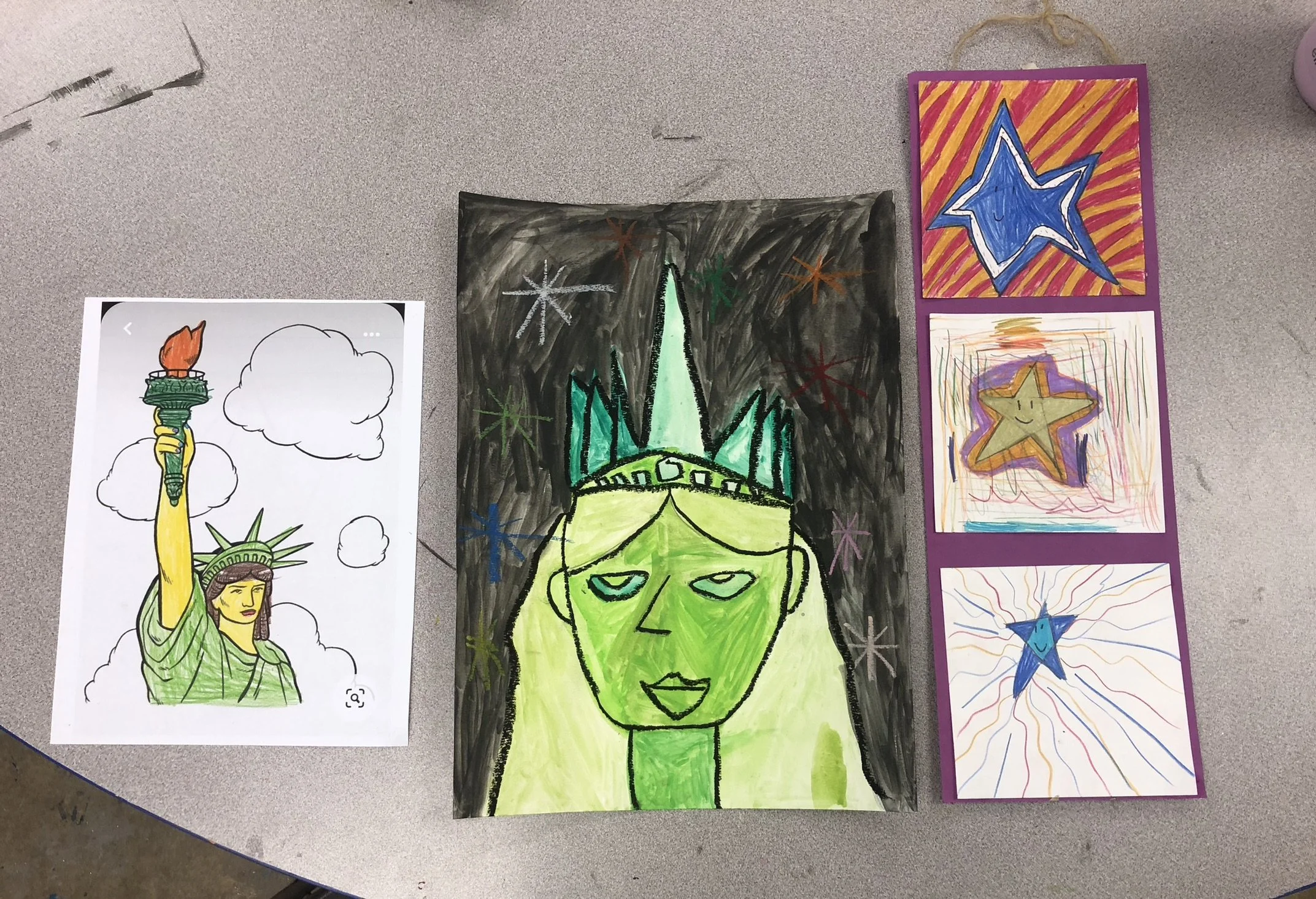 Collection of children's artwork featuring a drawing of the Statue of Liberty, a colorful drawing of a green face with a crown, and three star-themed cards with smiling stars in different colors and backgrounds, all displayed on a gray tabletop.