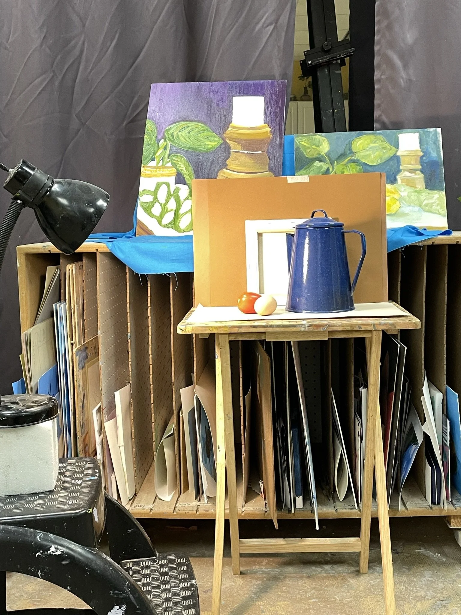 Art studio with completed paintings of vases and leaves on easels, a table with art supplies, and a bookshelf filled with art books and folders.