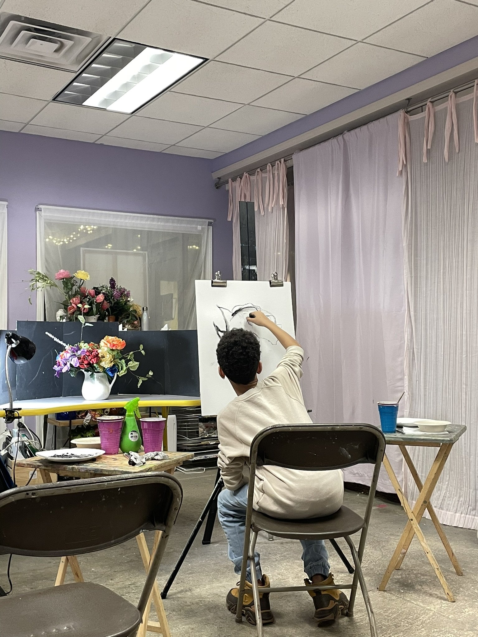 A person drawing on a white canvas in an art studio. The studio has purple walls, pink curtains, and various flowers and art supplies on tables.