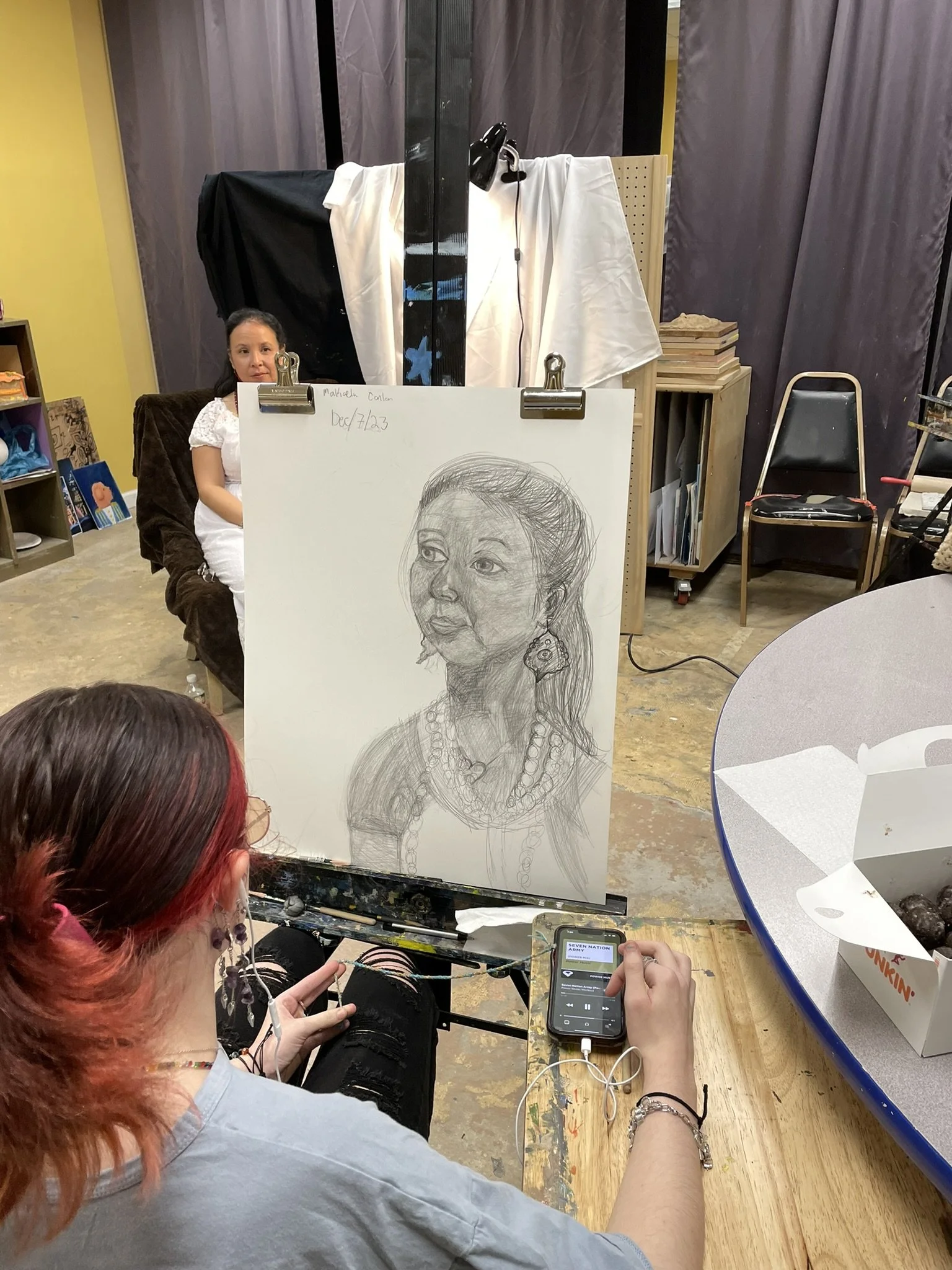 A woman with red hair and glasses listening to music and working on a drawing of a woman's portrait on a canvas. A young girl is seated on a chair in the background, and art supplies and a table with snacks are visible in the room.