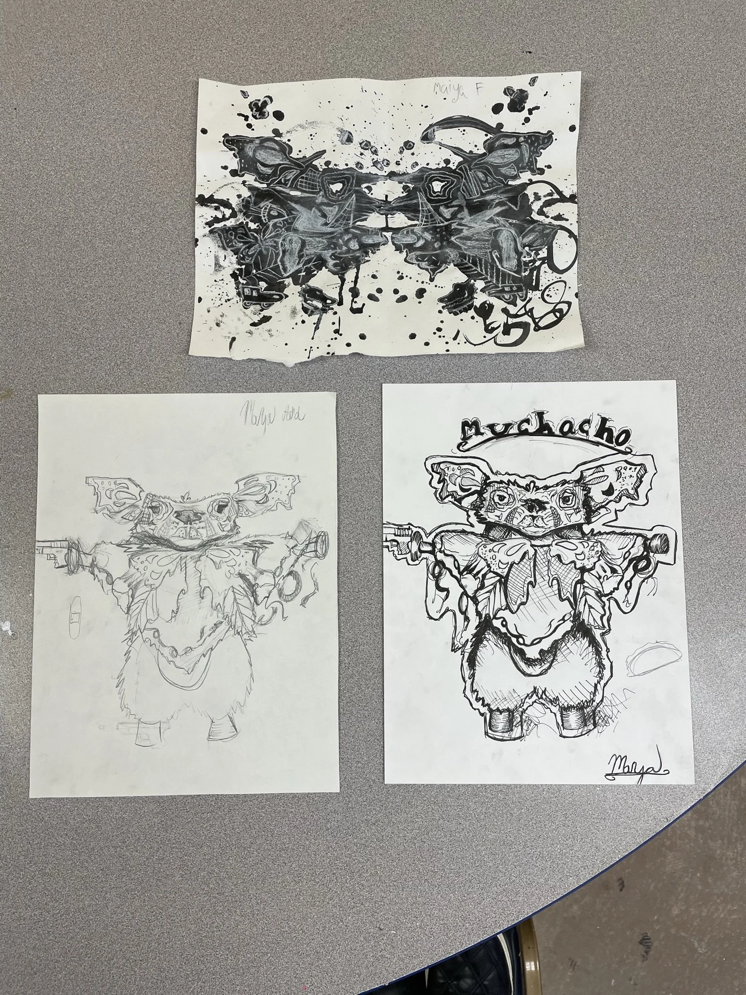 Three drawings of a butterfly with a pig's face, one colored with black ink and two sketches, one in pencil and one in ink, on white paper. The word "Mudcat" is written on the ink drawings. The drawings are placed on a gray table.