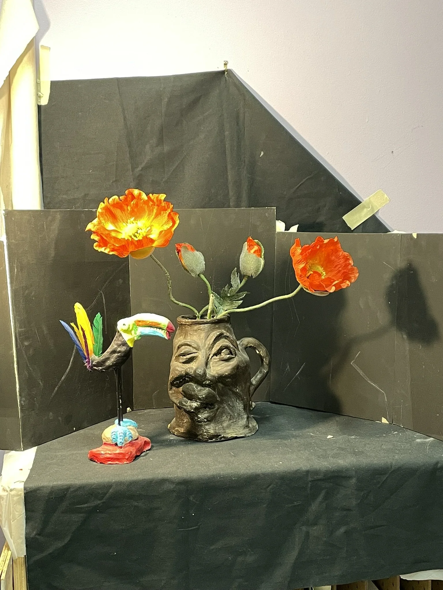 A black ceramic mug sculpture with a face holding a bouquet of orange poppy flowers on its top. A decorative toucan figurine stands to the left of the mug, with colorful feathers and a rainbow beak. Black panels form a backdrop behind the objects, pl