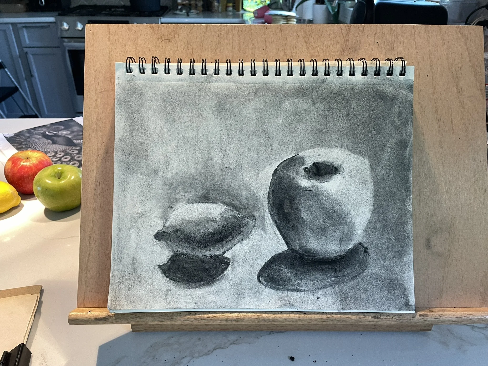 A grayscale watercolor painting of two apples, one whole and one cut open, with a smaller round object and shadows, on a wooden easel.