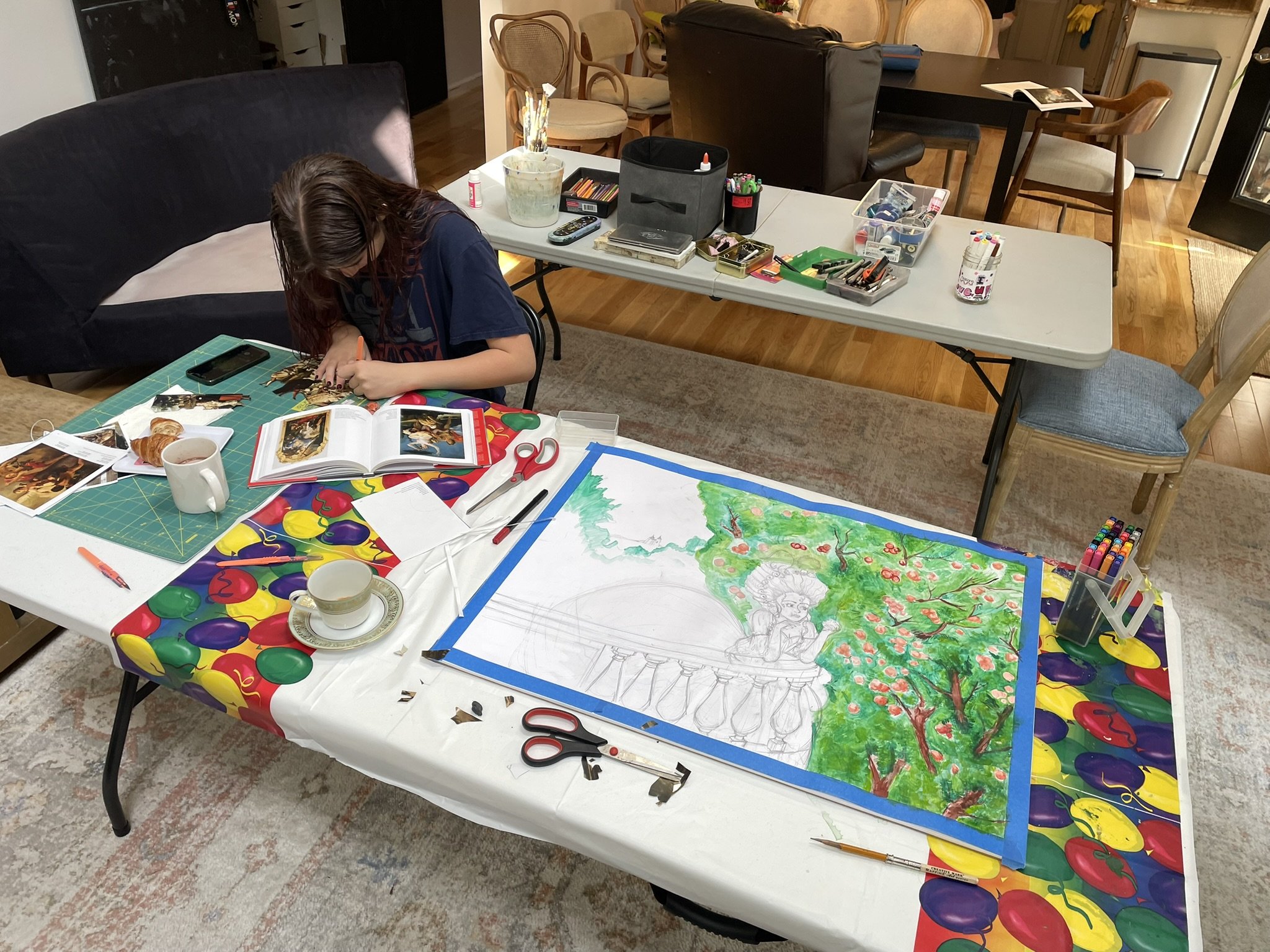 A young girl is working on an art project at a table covered with art supplies, photo prints, a cup of tea, and a book. She is painting a large watercolor piece of a scene with trees and a statue.
