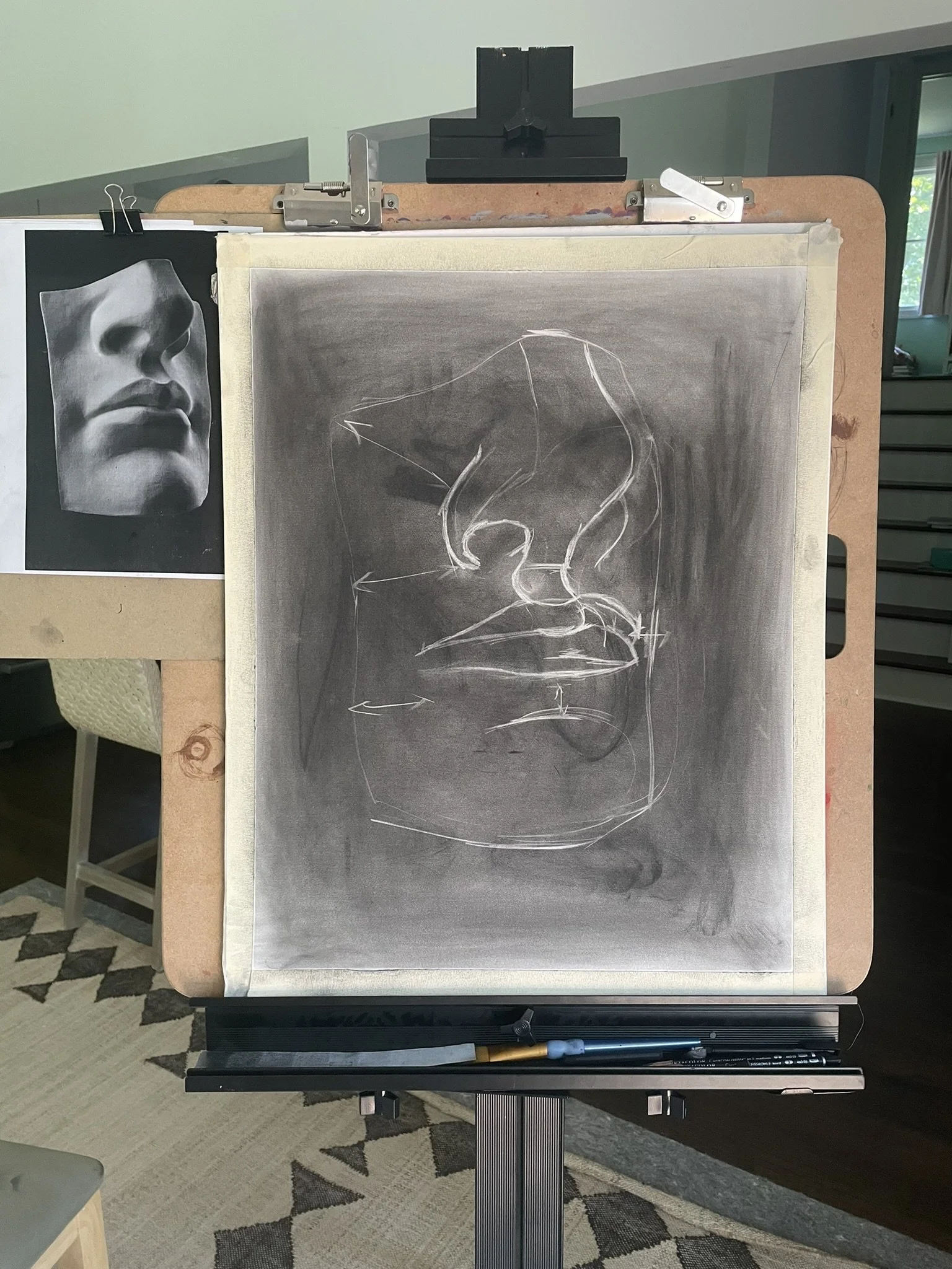A charcoal sketch of a human face in progress on an easel, with a reference photo of a mouth and nose on the side.